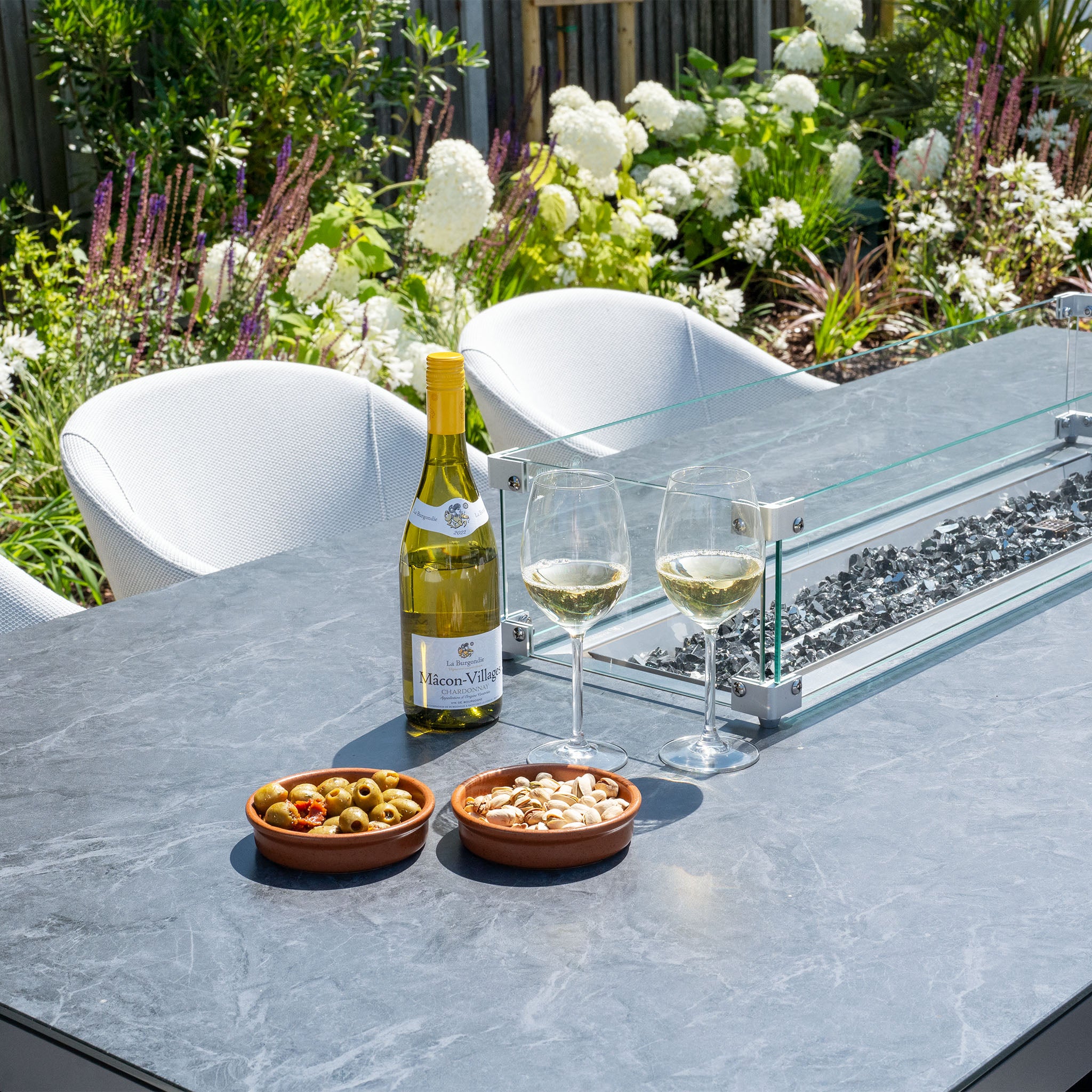 Outdoor table with two glasses of white wine, a wine bottle, bowls of olives and nuts, and a flower garden in the background, complemented by the Ali testing vimeo - low res featuring hydrophobic thin rope weave for a stylish touch.