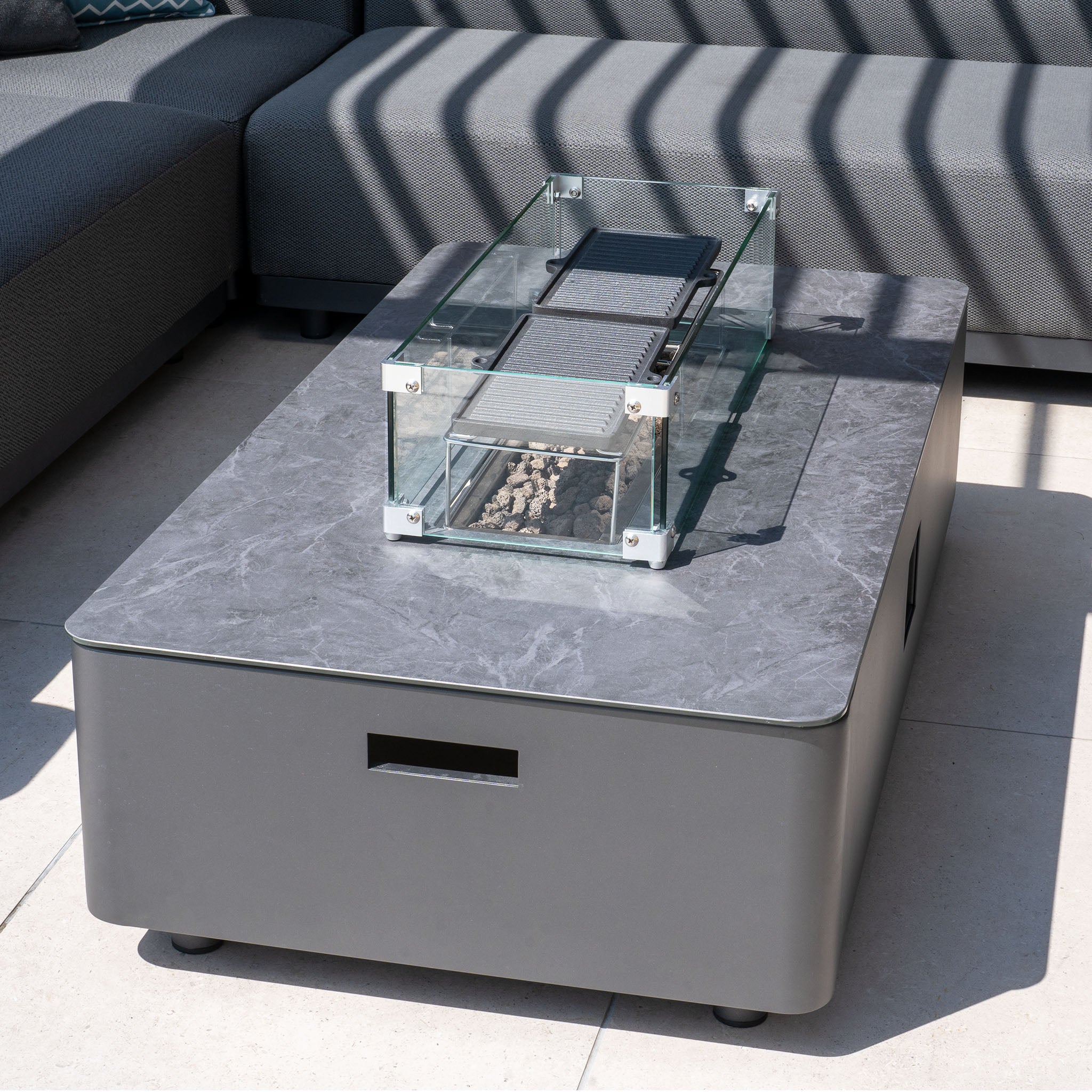 The Ceramic Firepit Coffee Table in Grey is a sleek, modern outdoor fire pit featuring a glass enclosure and an aluminium frame finished with TIGER Drylac® Powder Coating, set on a gray stone table on a sunlit patio.