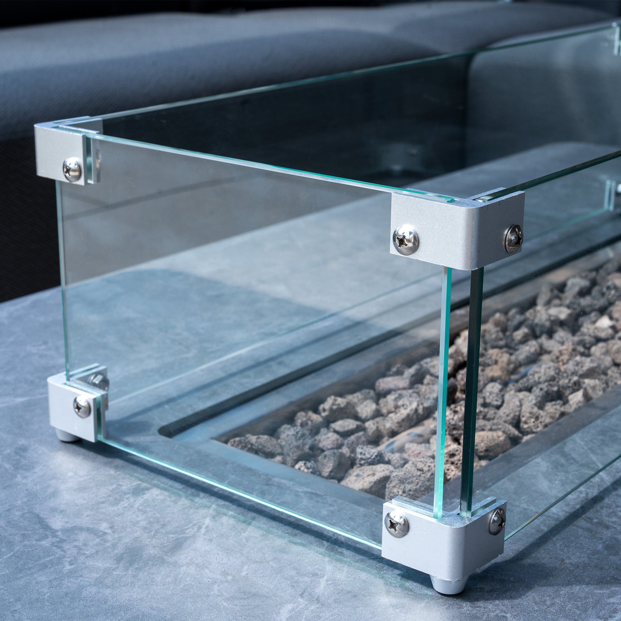 Close-up of a glass enclosure with metallic corner brackets, containing gray stones on the Ceramic Firepit Coffee Table in Grey with an Aluminum Frame on a gray marble surface.