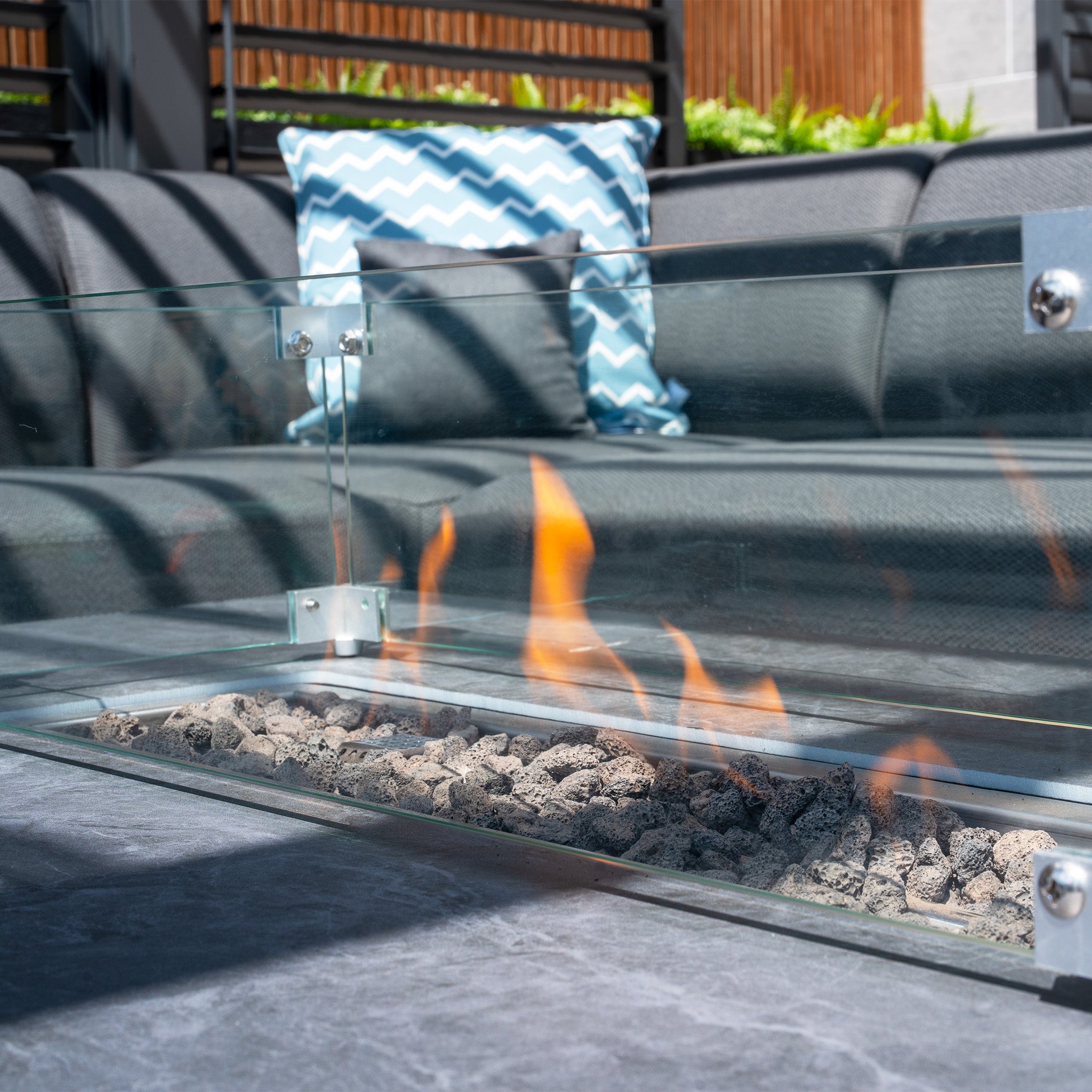 The Luna Outdoor Fabric Corner Sofa Set with Firepit Coffee Table in Grey features a glass-enclosed fire pit, flickering flames, and a grey couch adorned with a blue chevron-patterned Luna pillow, offering a maintenance-free experience.