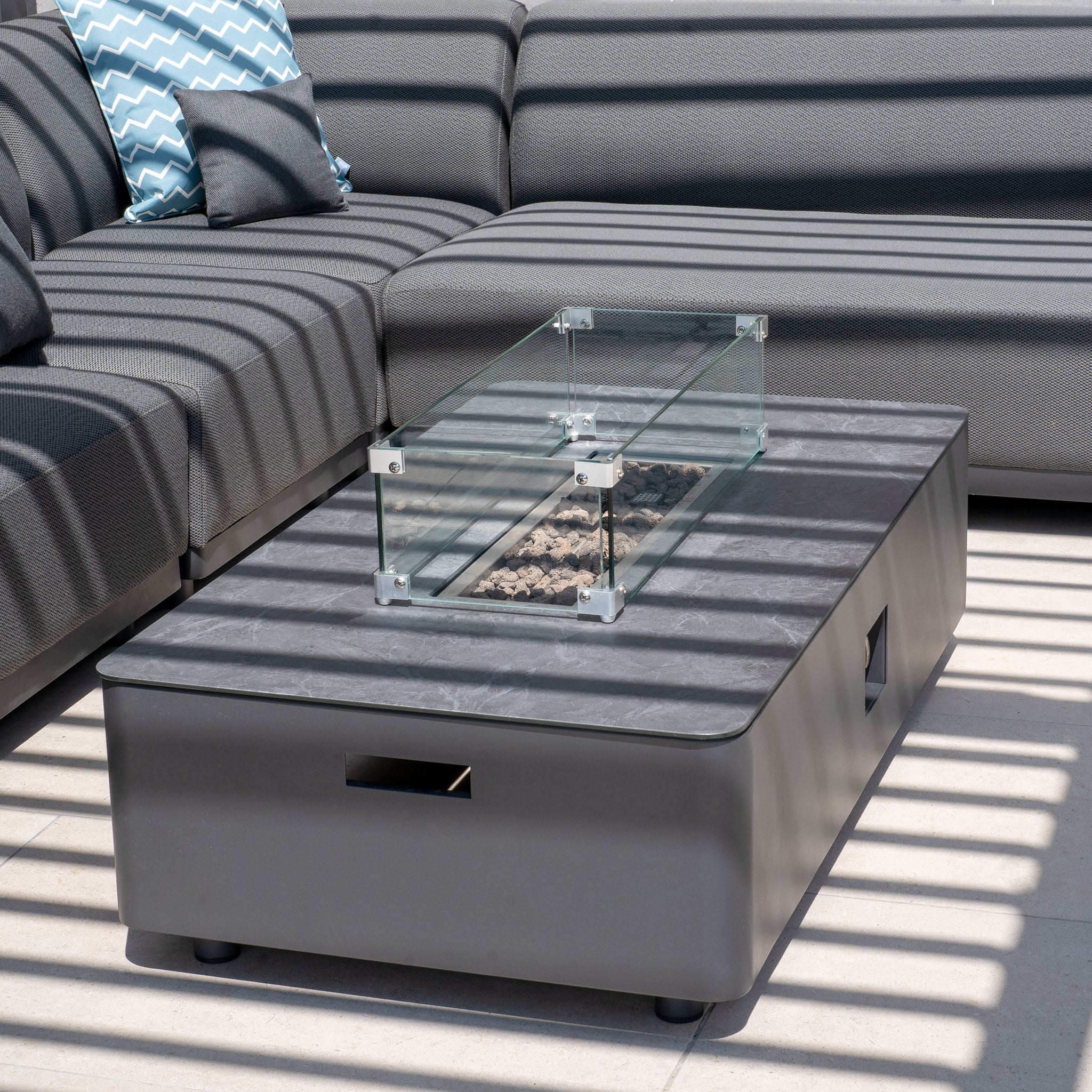 Modern outdoor patio with a black sectional sofa and a Ceramic Firepit Coffee Table in Grey under striped sunlight and shadow patterns. The aluminum frame of the furniture is finished with TIGER Drylac® Powder Coating for durability and style.