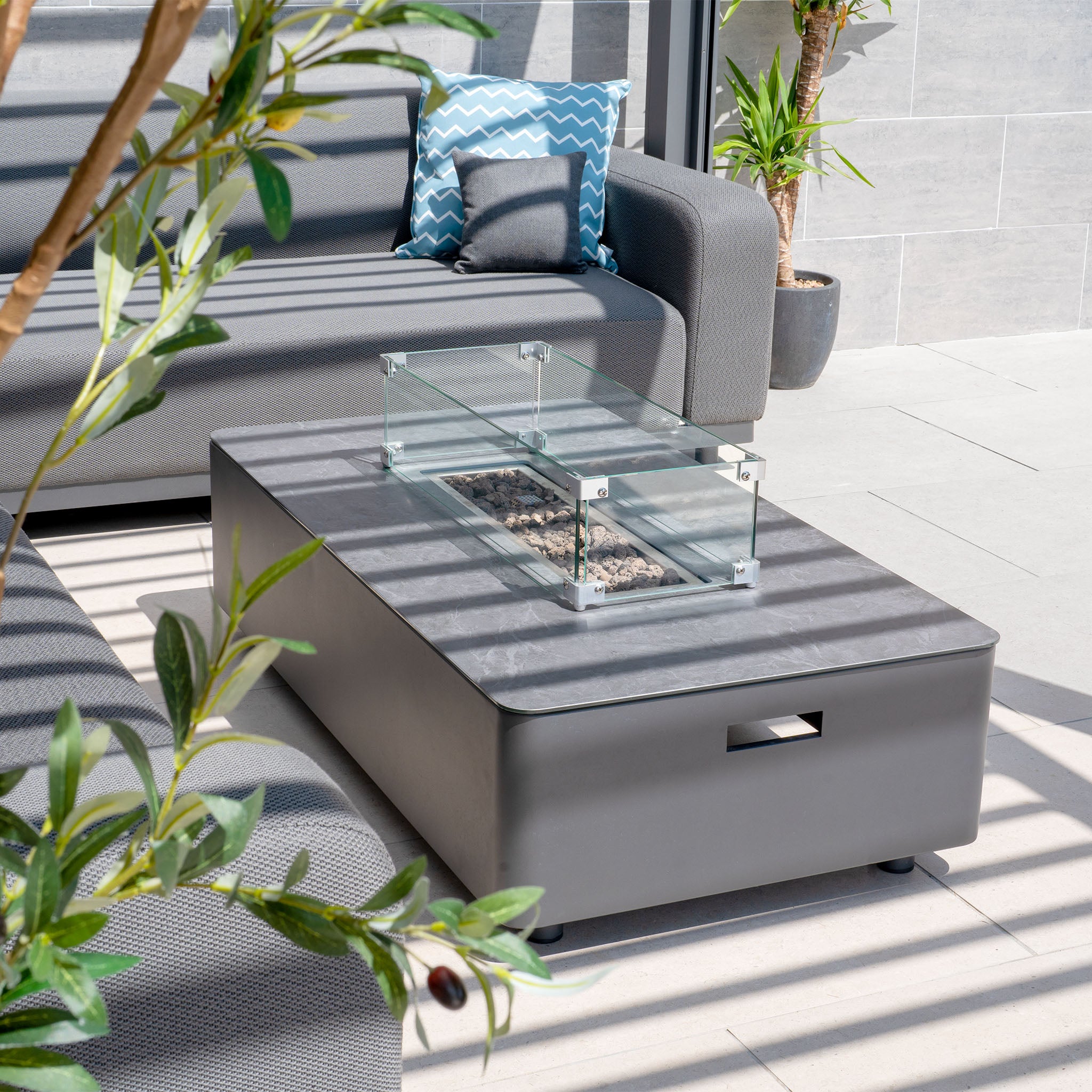 Modern outdoor patio featuring the Luna Outdoor Fabric Corner Sofa Set in grey with a maintenance-free firepit coffee table; enhanced with plants and geometric-patterned cushions.