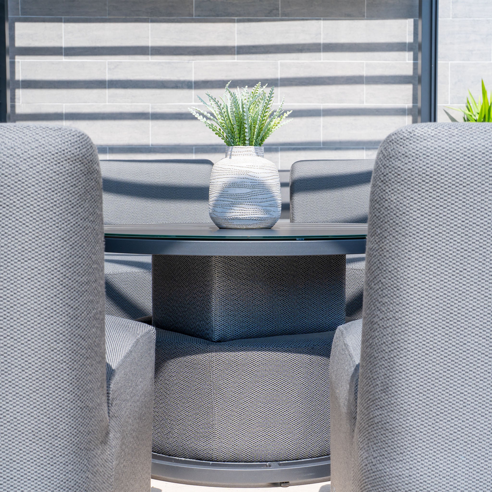 The Luna Outdoor Fabric Lifestyle Suite with 4 Grey Sofas surrounds a glass-top table adorned with a white vase containing green plants, all set against a modern gray wall.