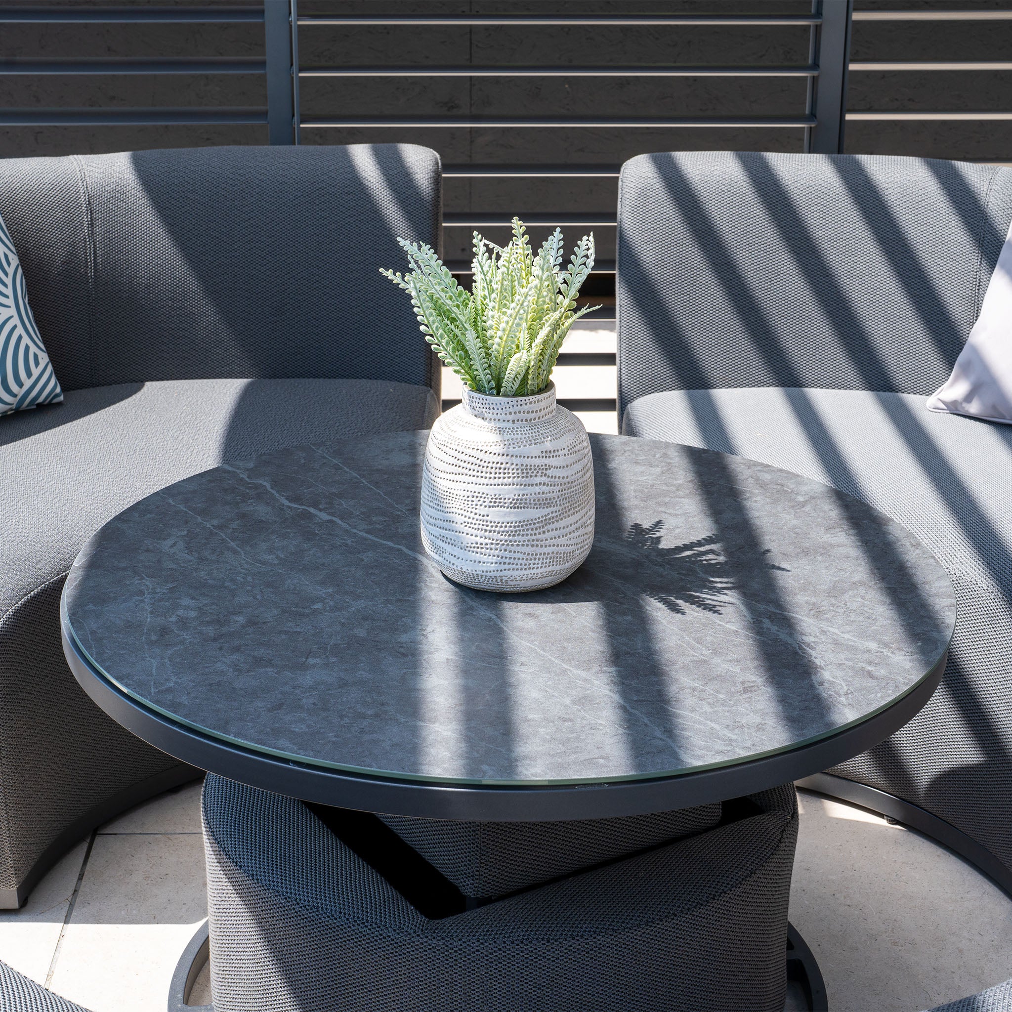 Modern patio setup with the Luna Outdoor Fabric Lifestyle Suite, featuring 4 grey upholstered sofas made from all-weather material under sunlight, a round table, and a potted plant centerpiece.