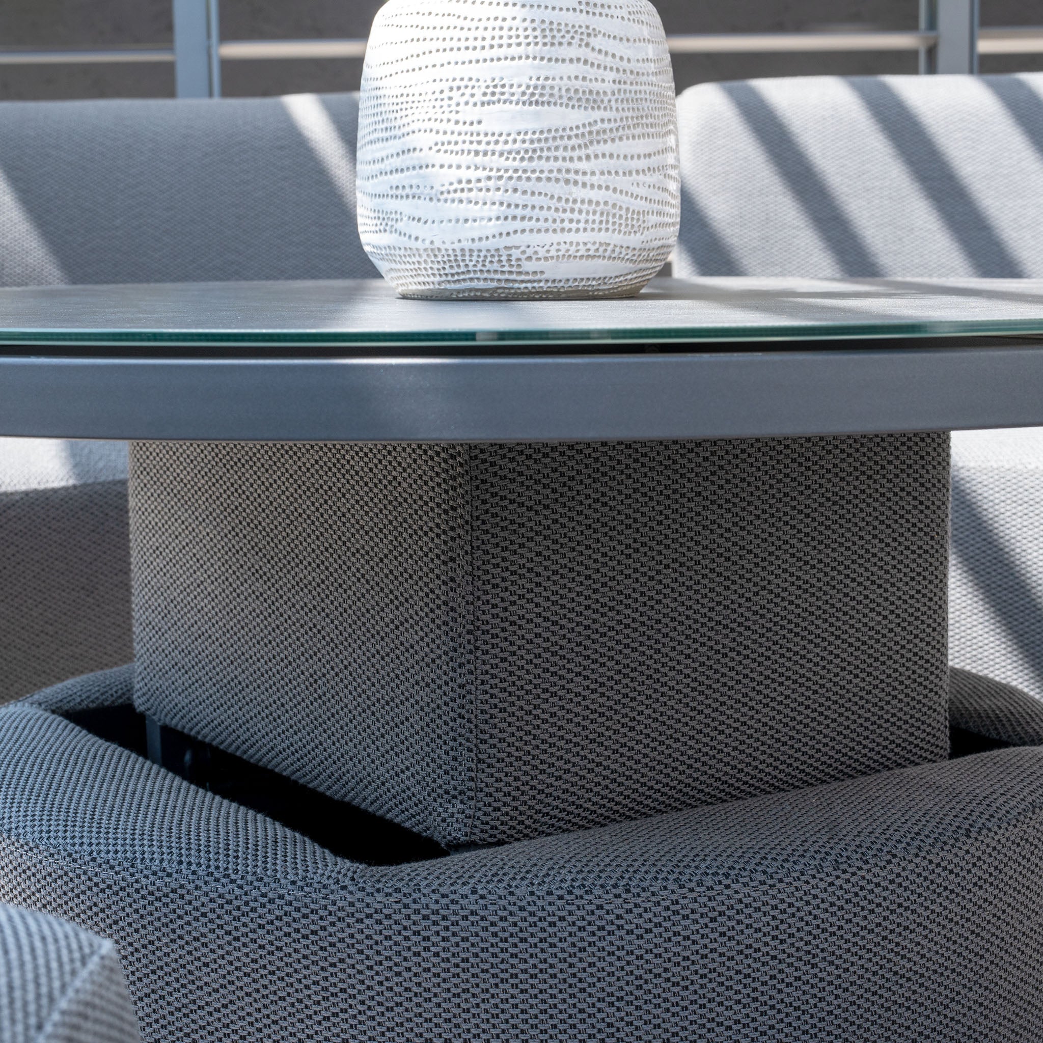 Close-up of a round glass table with padded seating in Luna Outdoor Fabric Lifestyle Suite with 4 Sofas in Grey, featuring a white textured vase on top, in a brightly lit room showcasing Harbour Lifestyle Luna's all-weather material.