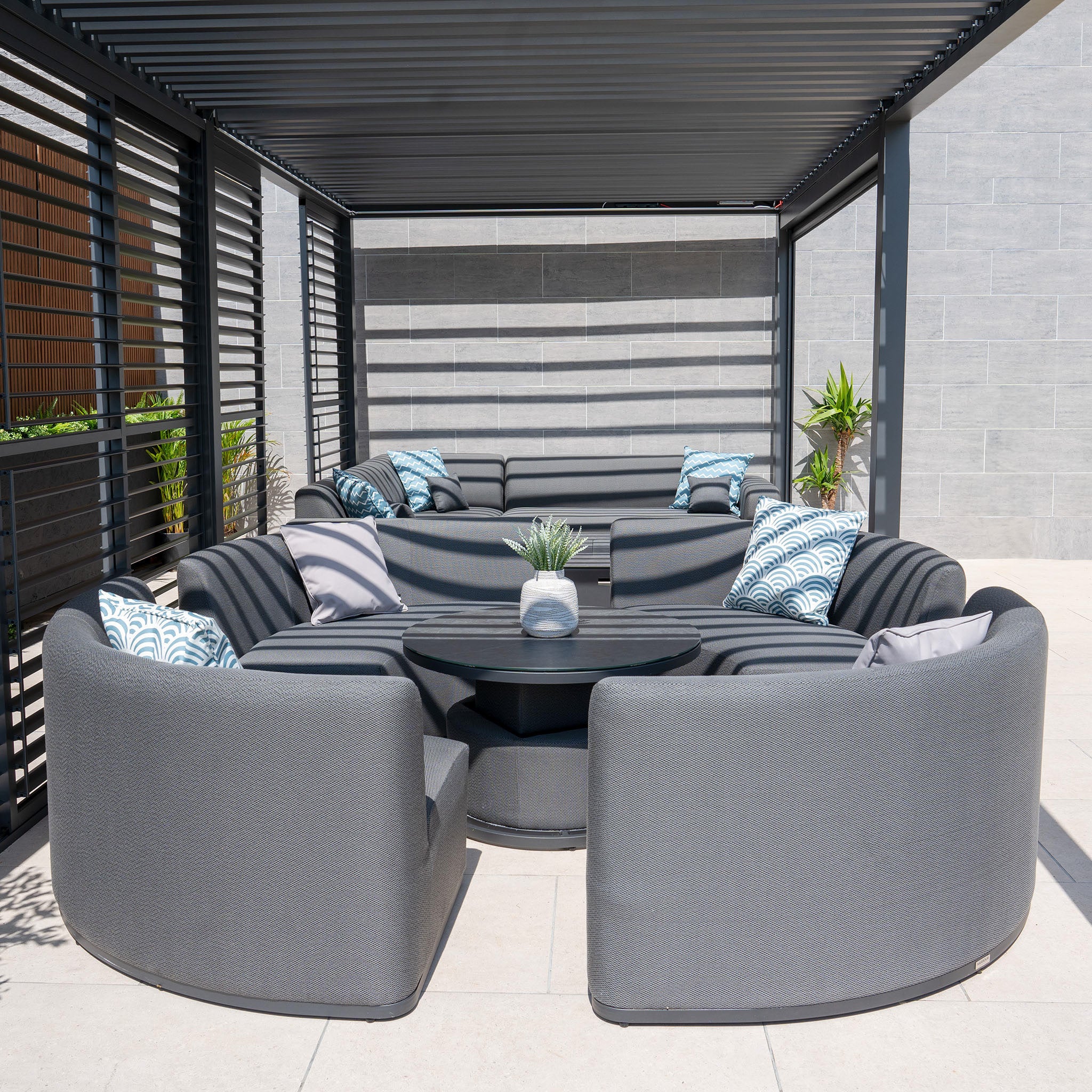 Modern outdoor seating area featuring the Luna Outdoor Fabric Lifestyle Suite with 4 Sofas in Grey by Harbour Lifestyle, accented with blue and white pillows, a coffee table adorned with a plant, and a pergola overhead. This setup is crafted from all-weather materials designed for durability and style.