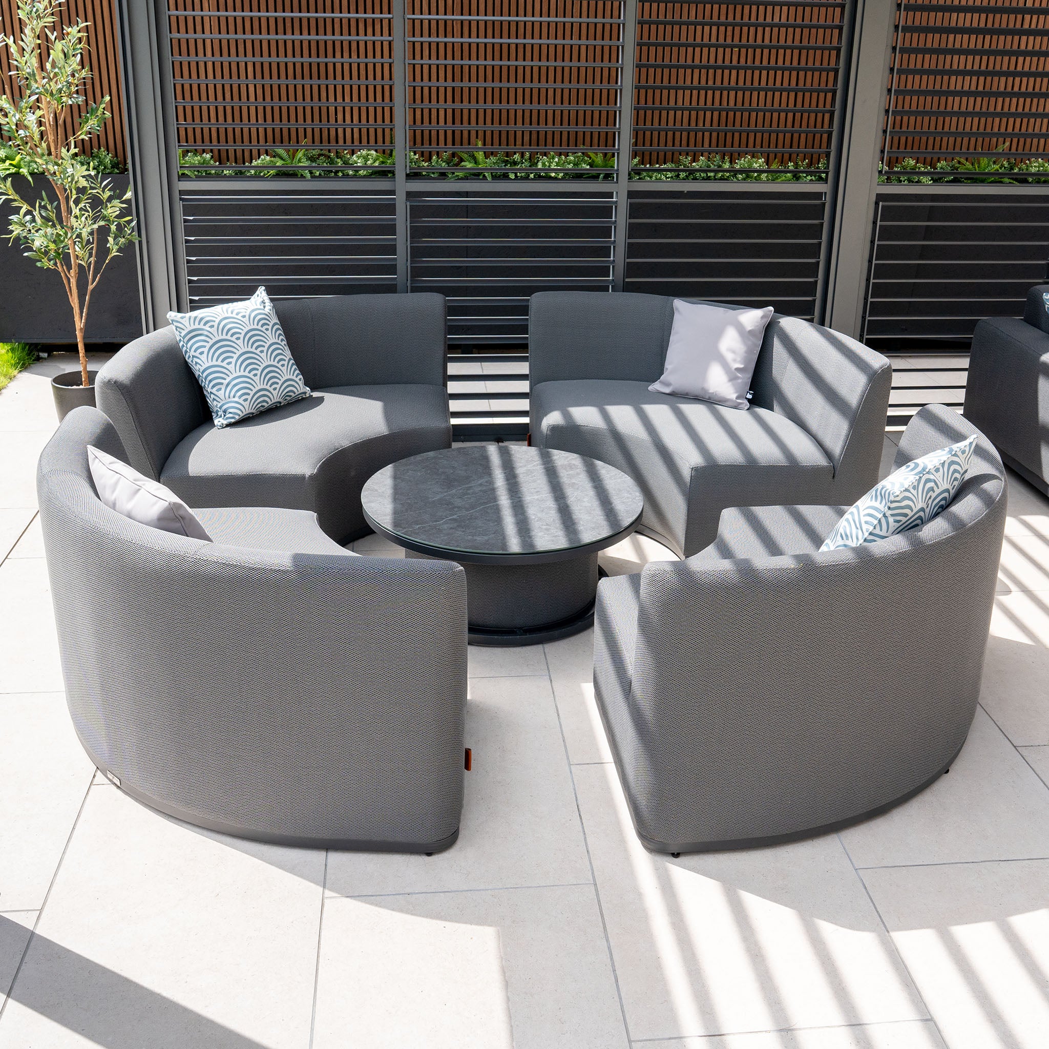 Outdoor seating area featuring the Luna Outdoor Fabric Lifestyle Suite, consisting of four grey cushioned sofas arranged around a round black table on a tiled patio.