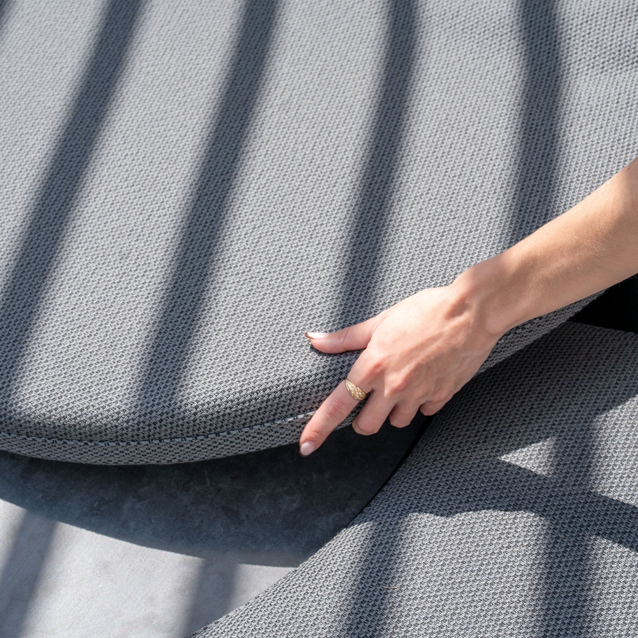 A hand adjusts a round, textured gray cushion made from the Luna Outdoor Fabric Lifestyle Suite with 4 Sofas in Grey under shadowed bars on a light surface.