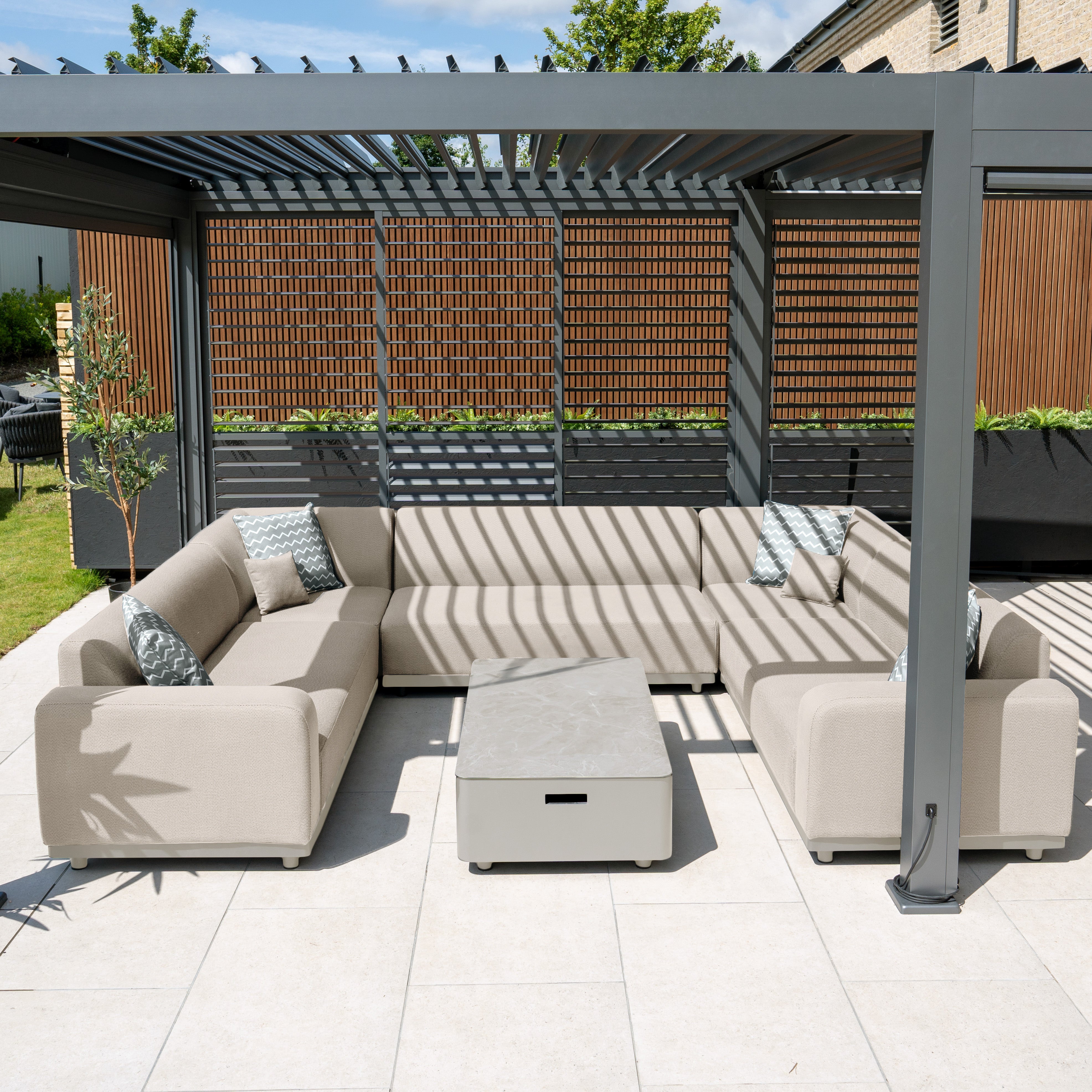 The Luna U-Shape Outdoor Fabric Sofa Set with Coffee Table in Fawn provides modern outdoor seating beneath a pergola, creating textured shadows. It features weather-proof outdoor fabric and quick-dry foam cushions, complemented by lush greenery and a wooden privacy screen for added serenity.