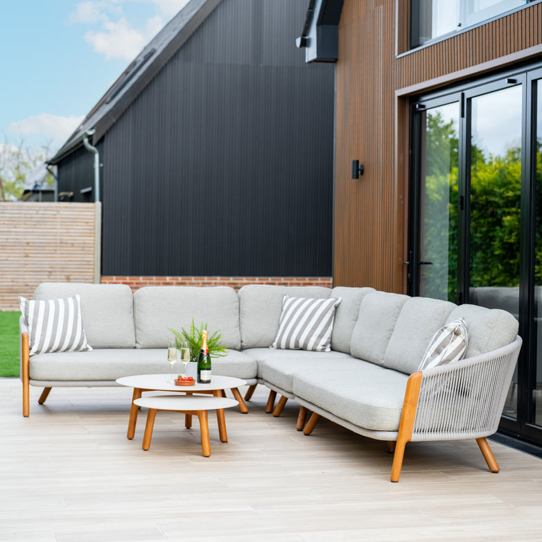 Discover the Merano Large Corner Group Set with Coffee Table in Latte in this outdoor living space, showcasing a cozy sectional sofa with striped cushions and an elegant round table accented by refreshing drinks and a stylish plant, all highlighted by an exquisite olefin rope weave design.