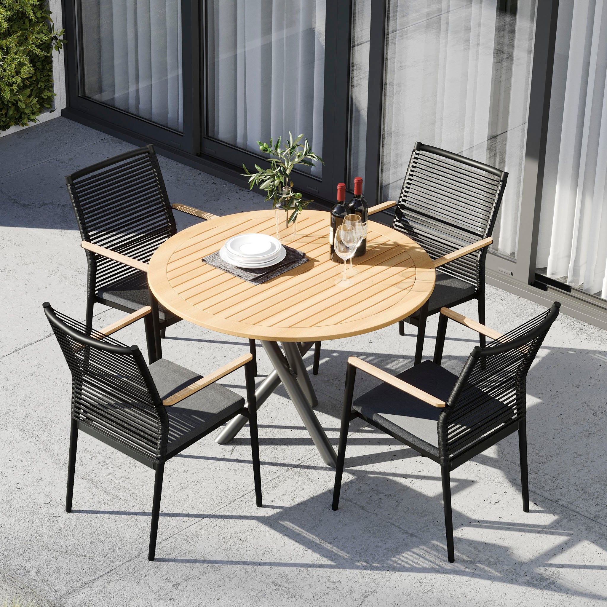 Outdoor patio featuring the Portland 4 Seat Round Dining Set with a teak table in charcoal, complemented by four flat rope weaved armchairs, along with wine bottles, glasses, and plates on a concrete surface.