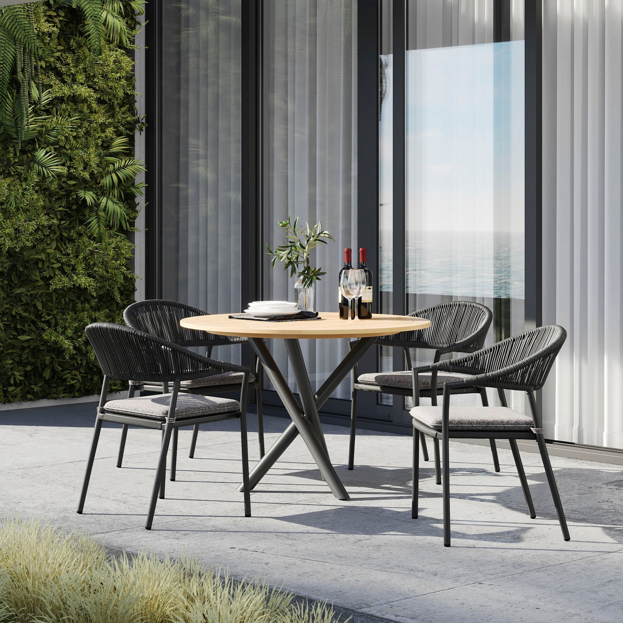 Outdoor patio featuring the Cloverly 4 Seat Round Dining Set with a teak table top in charcoal, adorned with wine bottles and glasses, near glass windows.