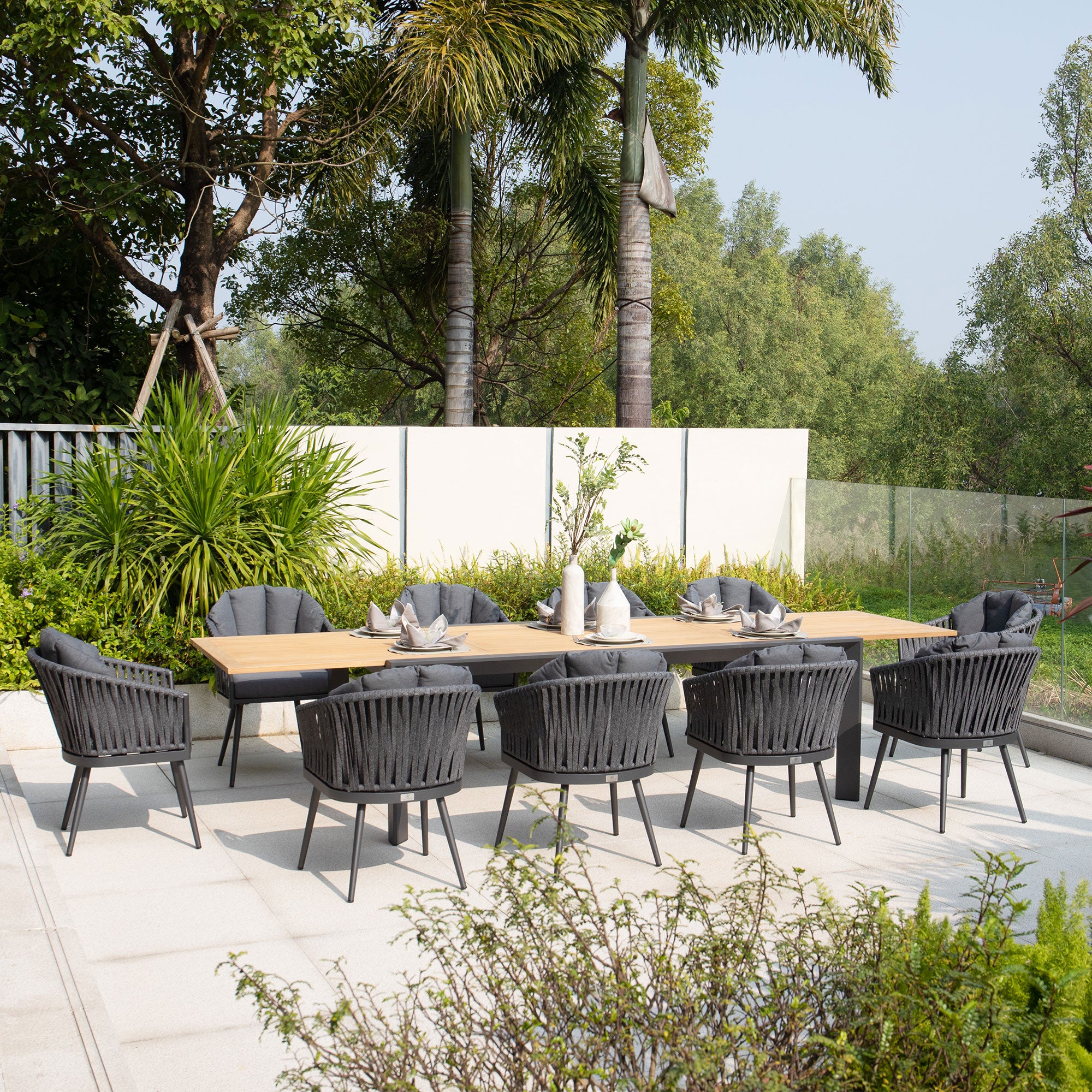 Outdoor patio with the Monterrey 10 Seat Rope Extending Teak Dining Set in Grey, featuring a teak extending table surrounded by ten Monterrey chairs with outdoor rope fabric, set against lush greenery and trees.