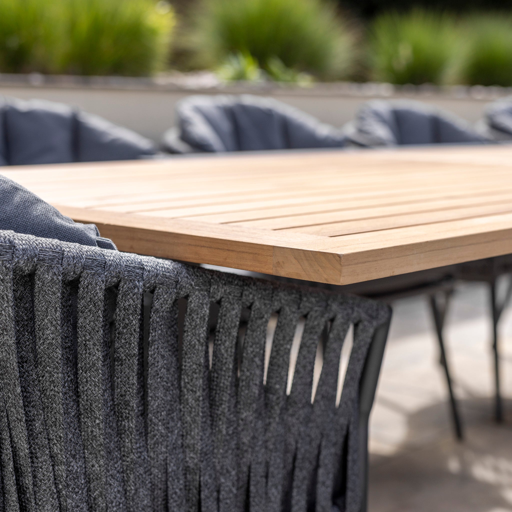 Close-up of the Monterrey 10 Seat Rope Extending Teak Dining Set in Grey, surrounded by green plants blurred in the background.