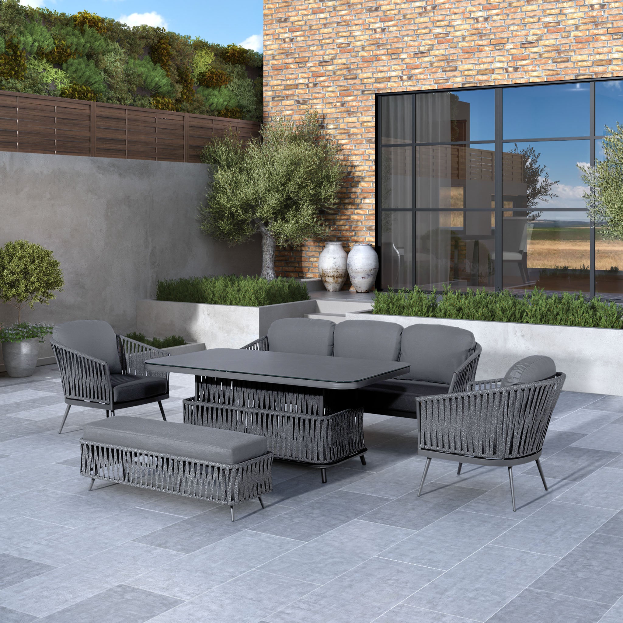 An outdoor patio featuring the Monterrey 3 Seat Rope Sofa Set with Rising Table in Grey, which includes a modern grey sofa, chairs, bench, and table with reflex foam fillings and a hydrophobic thin rope weave design, situated next to a brick building.