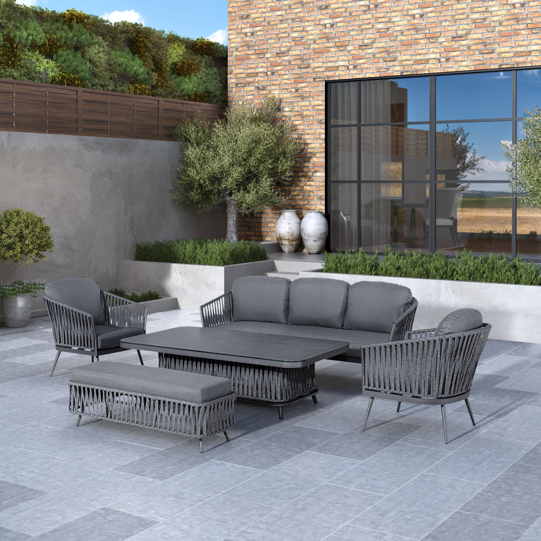 Modern outdoor patio featuring the Monterrey 3 Seat Rope Sofa Set with Rising Table in Grey, including a sofa and chairs with reflex foam fillings, placed against a brick wall.