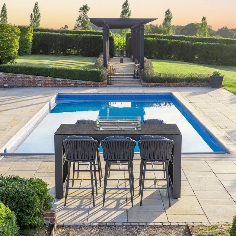 A modern backyard features a pool, three barstools from the Monterrey 6 Seat Rope Ceramic Firepit Bar Set in Grey at a high table made with hydrophobic thin rope weave, and a trellis pathway, set against a sunset sky.