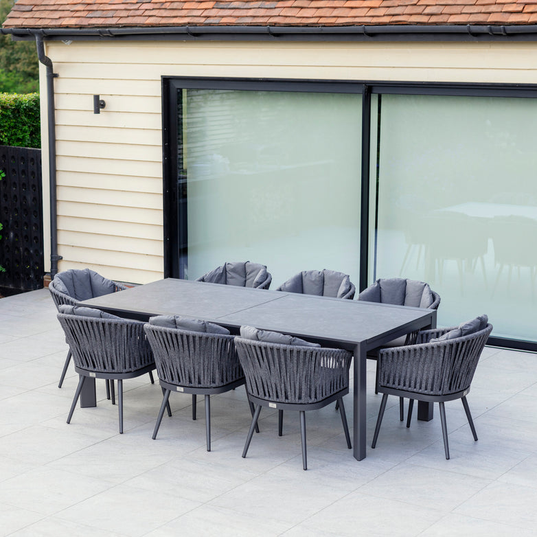 The Monterrey 8 Seat Rope Extending Dining Set in Grey, featuring a rectangular table and eight cushioned chairs adorned with outdoor rope fabric, is elegantly placed on a tiled patio next to a house with large windows.