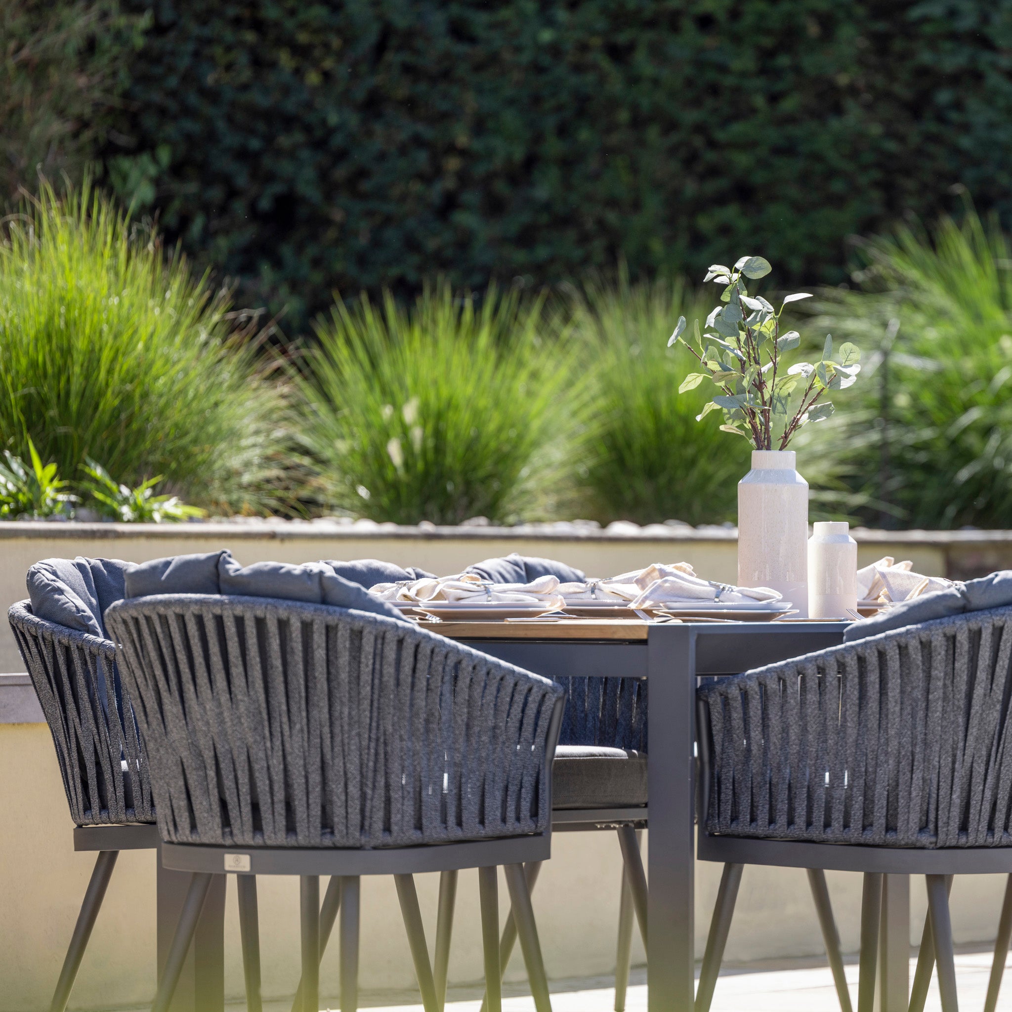 The Monterrey 10 Seat Rope Extending Teak Dining Set in Grey features cushioned seats and Outdoor Rope Fabric detailing. A vase with green branches adds a touch of elegance against the backdrop of lush green foliage.