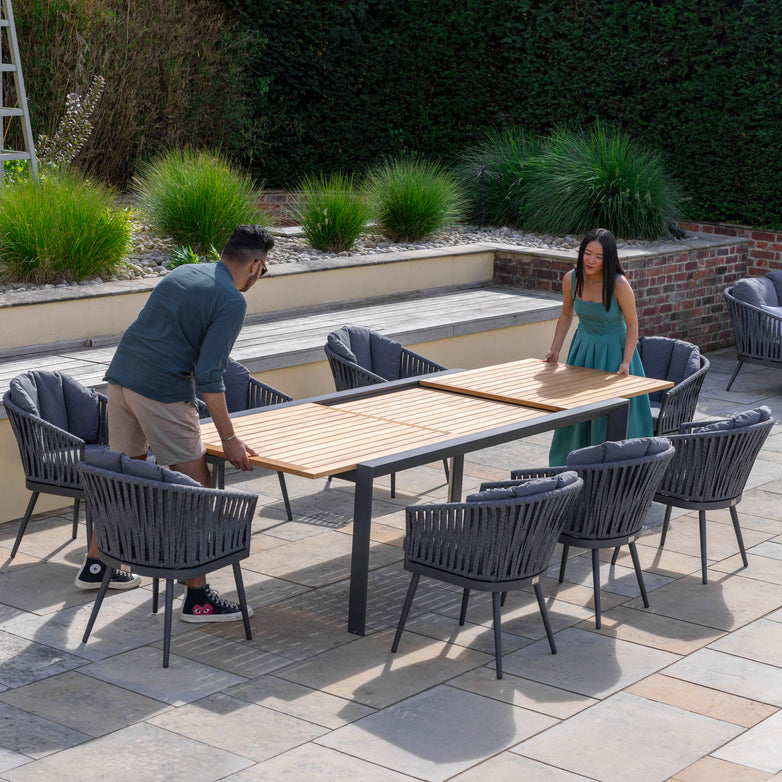 Two people adjust the Monterrey 10 Seat Rope Extending Teak Dining Set in Grey on a patio, surrounded by greenery.