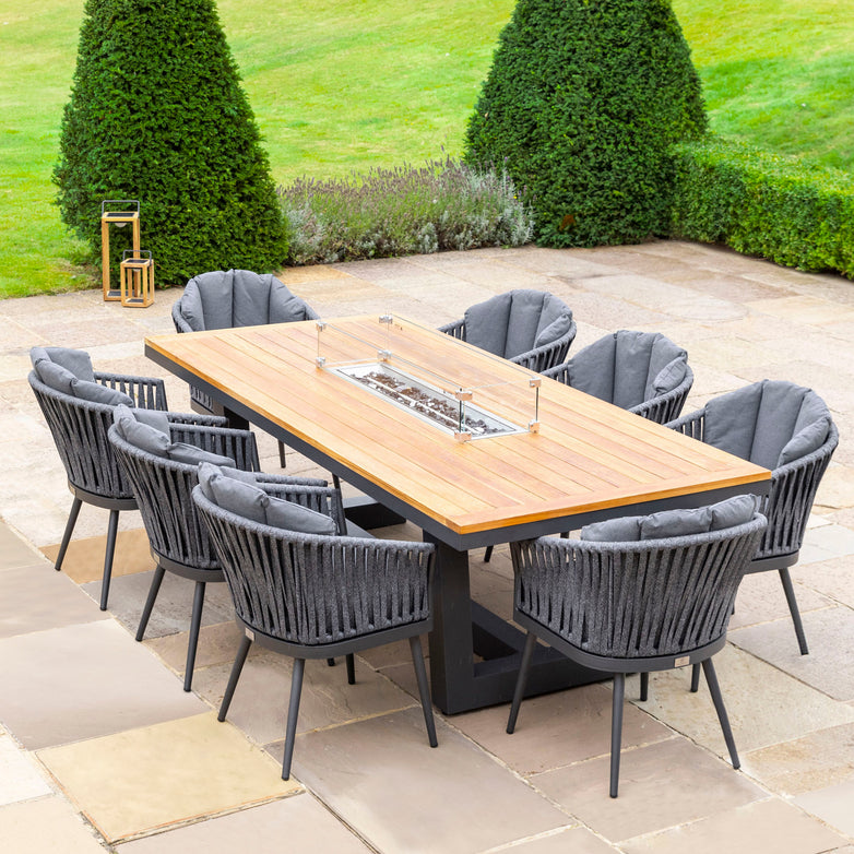 Enjoy your outdoor patio with the Monterrey 8 Seat Rope Rectangular Teak Firepit Dining Set in Grey: a stylish wooden table featuring a built-in fire pit and eight cushioned chairs, all enhanced by durable outdoor rope fabric and surrounded by lush greenery.