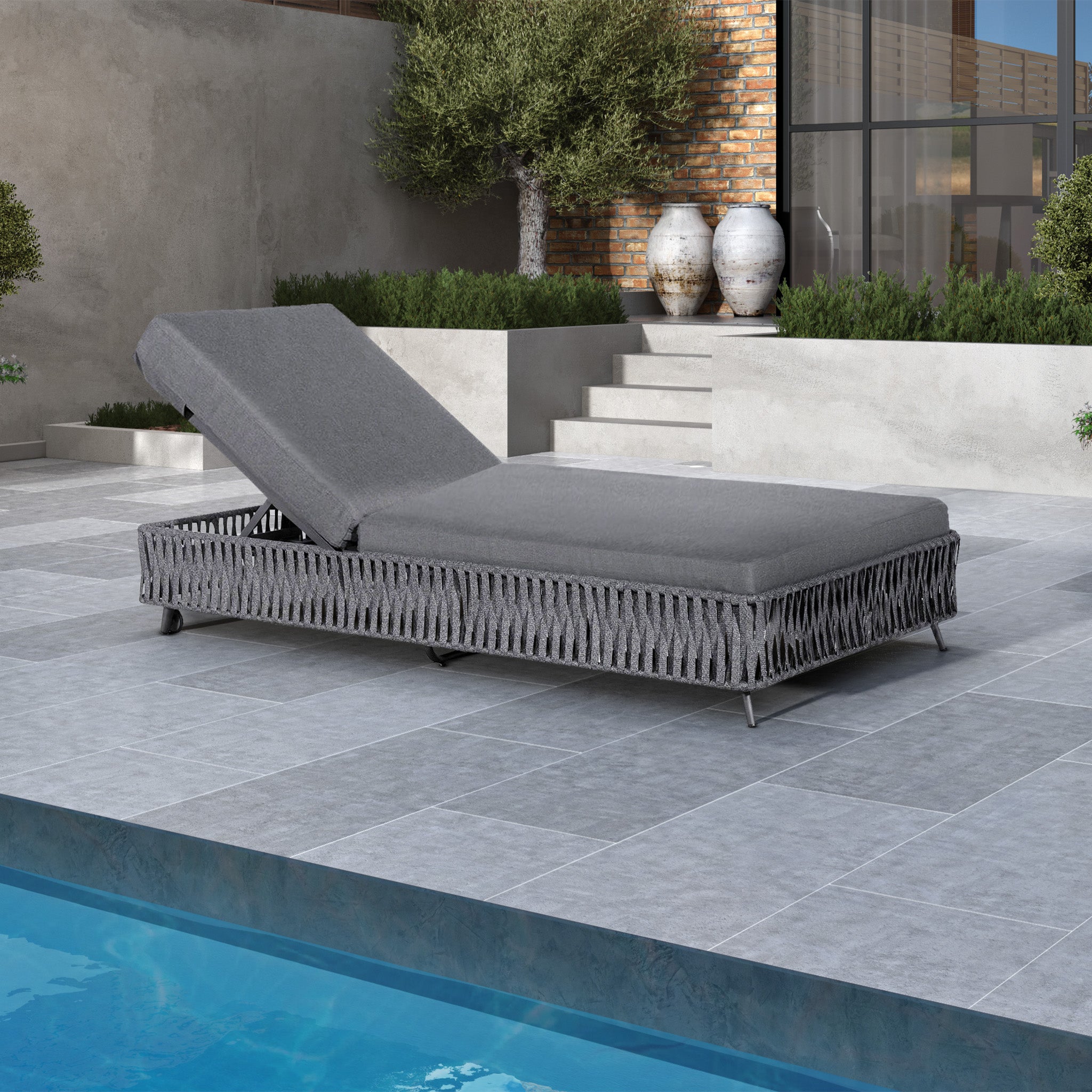 Monterrey Rope Sun Lounger in Grey by a poolside, set on a tiled patio with greenery and brick wall in the background, featuring durable Olefin fabric and reflex foam cushioning.