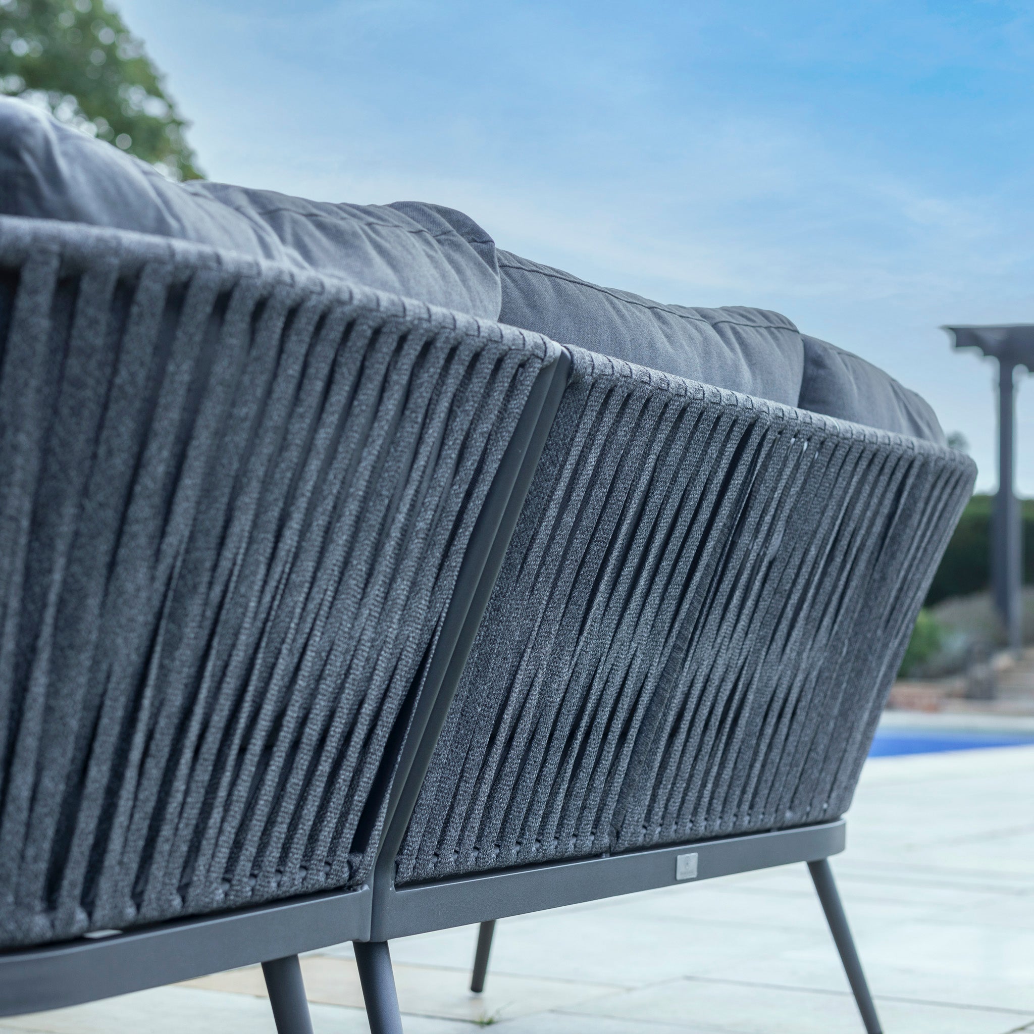 Close-up of a Harbour Lifestyle Monterrey U-Shape Rope Corner Dining Set with Rising Table in Grey, set on a patio with a view of the sky and a pergola.