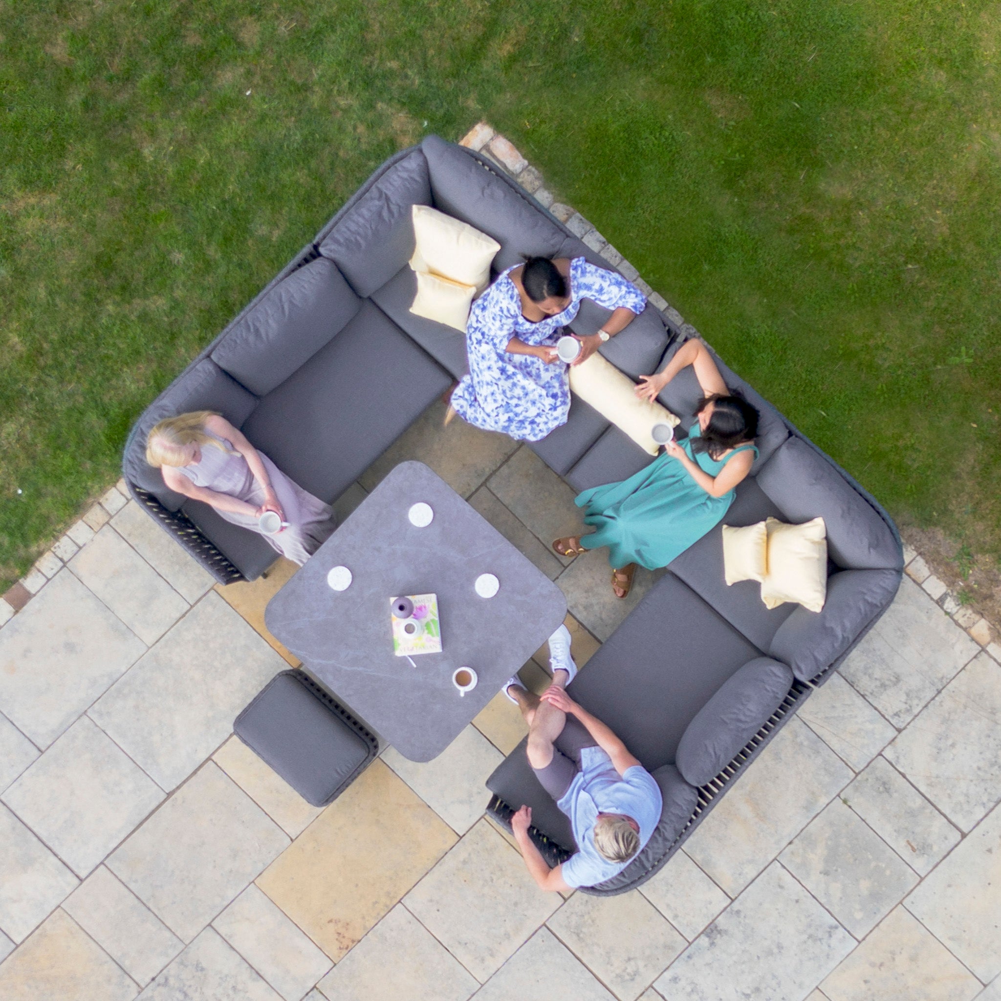 Four people sit around a Monterrey U-Shape Rope Corner Dining Set with Rising Table in Grey on outdoor patio furniture, viewed from above, with grass bordering the patio area.