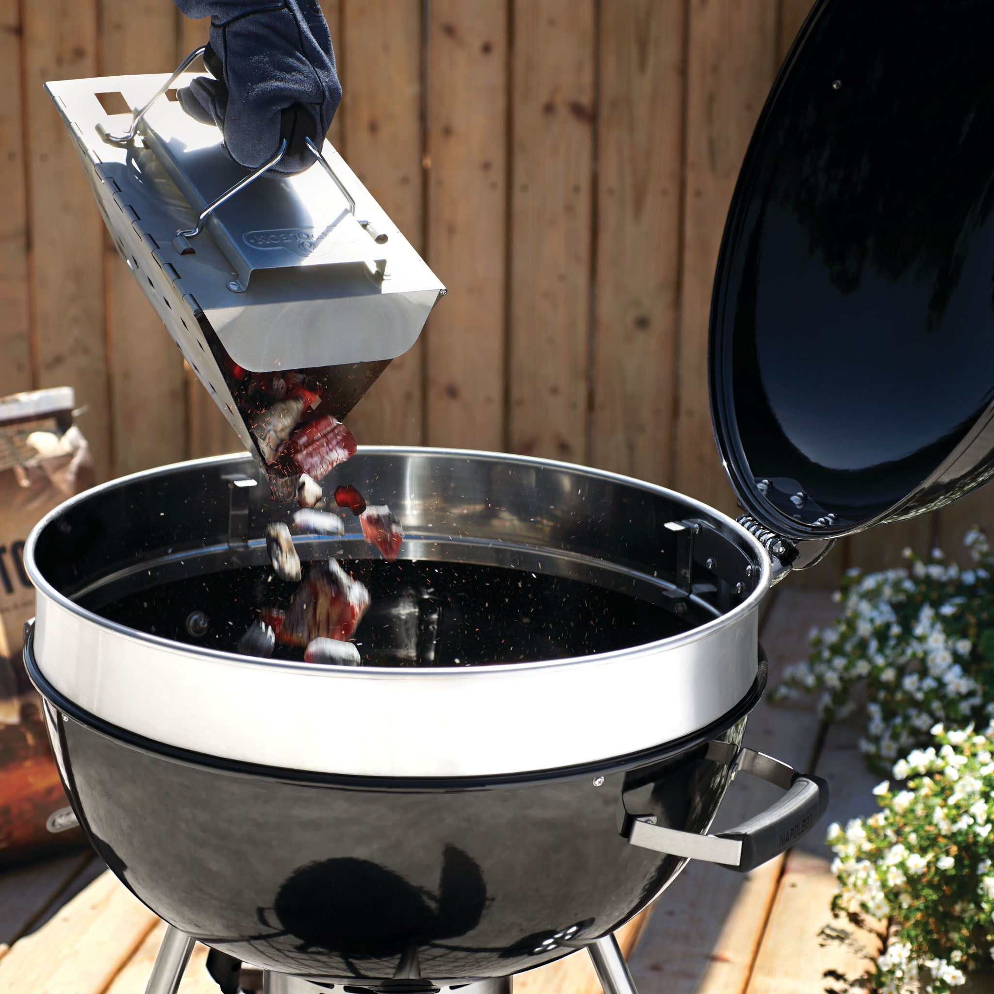 Someone is emptying a chimney starter of hot coals into a Napoleon Pro Charcoal Kettle Grill on a wooden deck, preparing for precise temperature control.