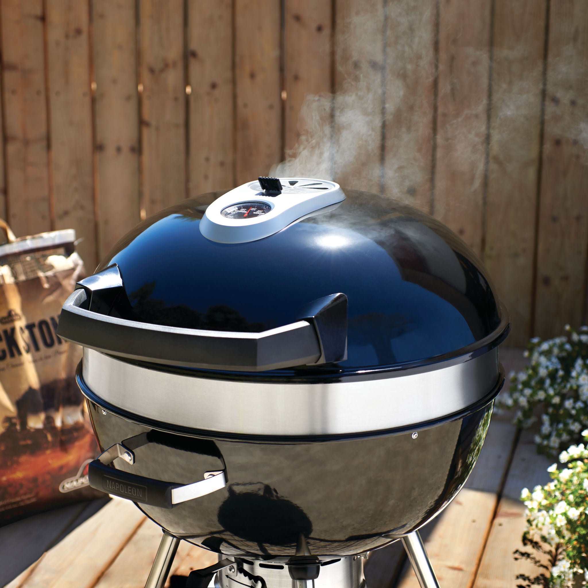 A black Napoleon Pro Charcoal Kettle Grill with smoke rising from it stands outdoors on a wooden deck near a wooden fence and a bag of charcoal, offering precise temperature control.