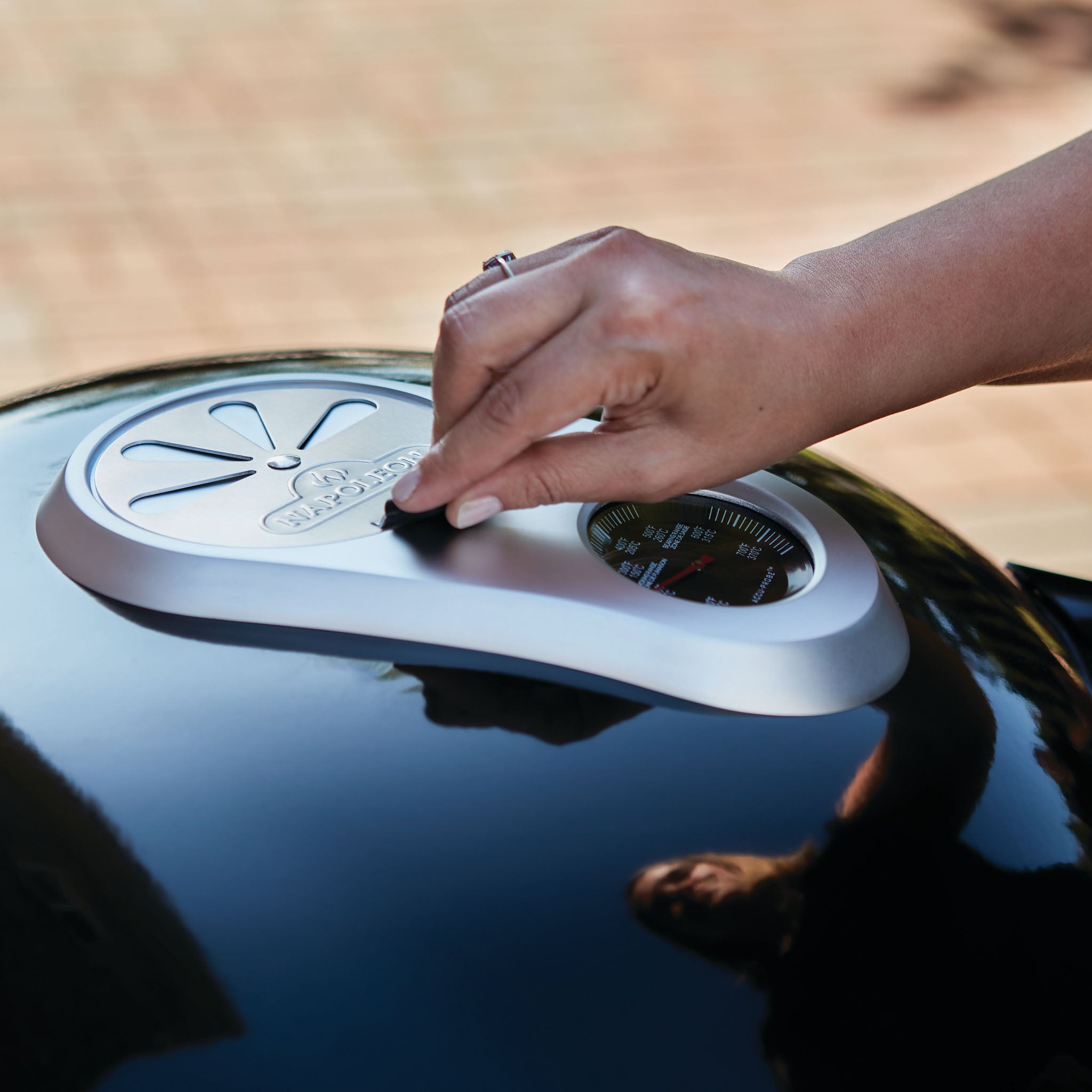 Close-up of a hand adjusting a dial on the lid of a gleaming black Napoleon Pro Charcoal Kettle Grill. Reflections on the grill surface highlight its precise temperature control features.