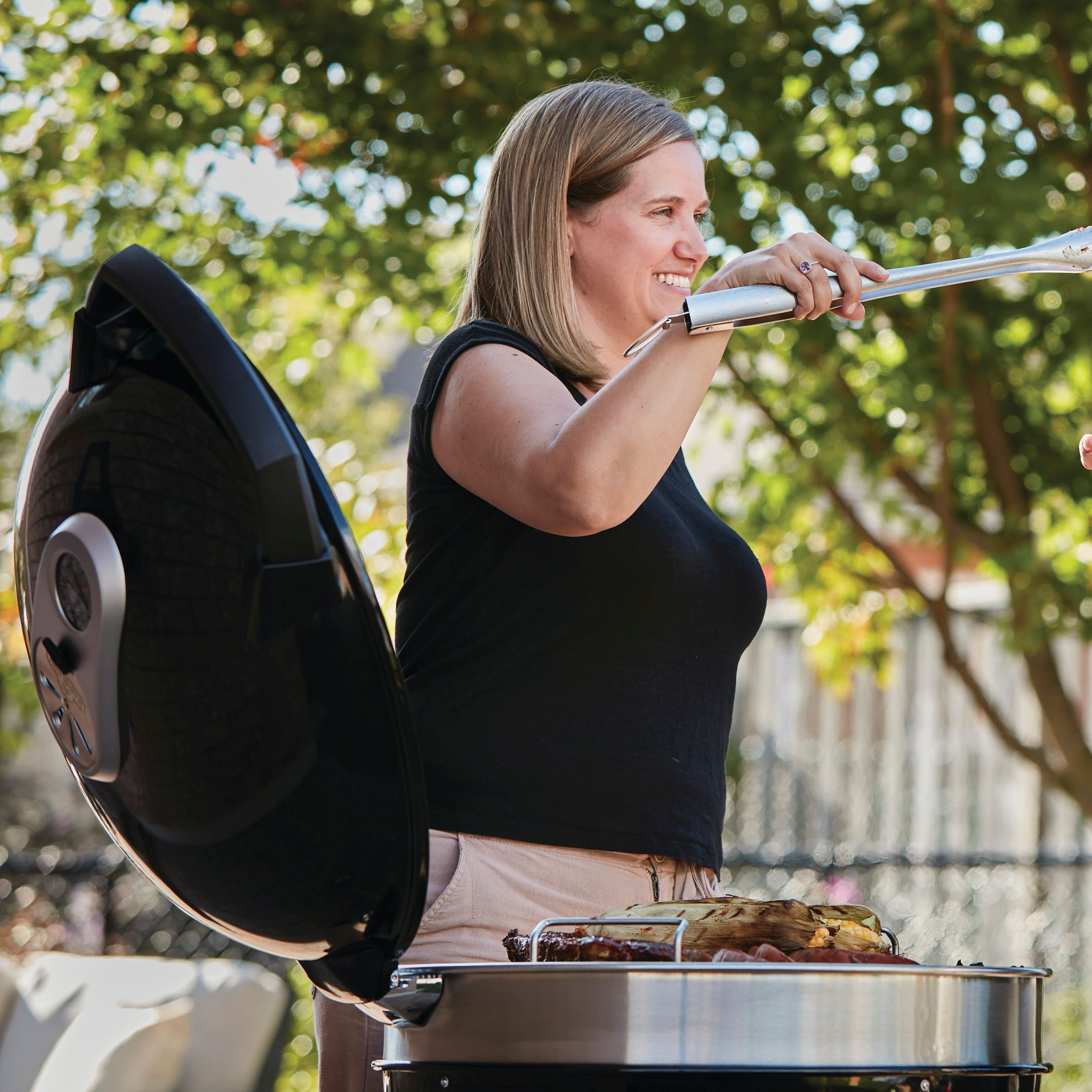 A smiling woman uses tongs to grill food on an outdoor Napoleon Pro Charcoal Kettle Grill, enjoying precise temperature control; trees and sunlight in the background.