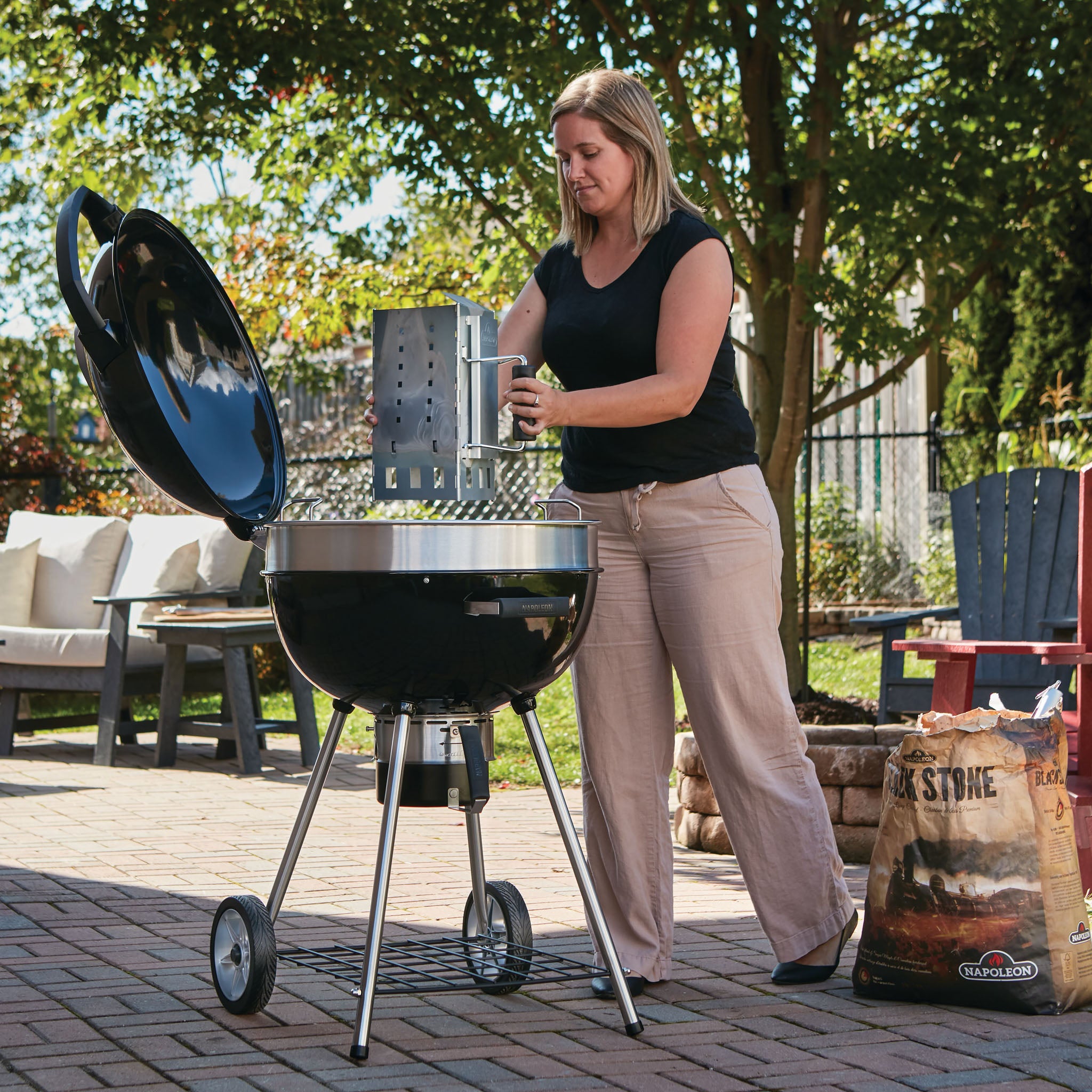 A woman, dressed casually, is setting up the Napoleon Pro Charcoal Kettle Grill outdoors on a sunny day. Patio furniture in the background adds to the ambiance as she enjoys the grill's precise temperature control.