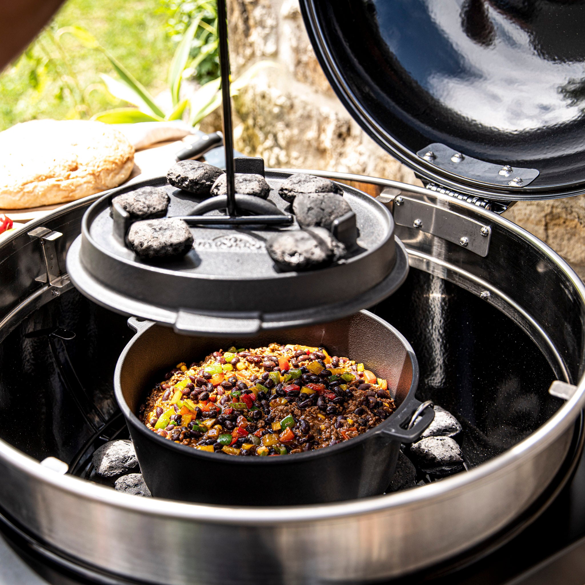 Cast iron pot with colorful stew, surrounded by charcoal briquettes, cooking inside a Napoleon Pro Charcoal Kettle Grill with an open lid.