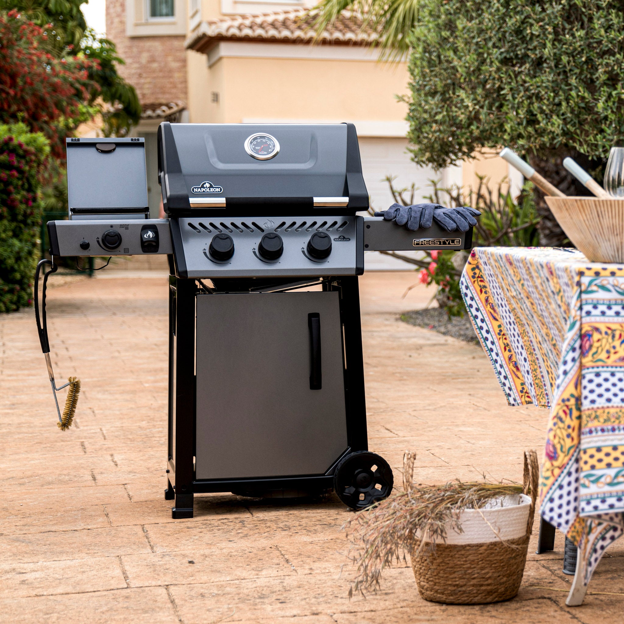 A black grill in an outdoor setting features gloves on a side shelf. A colorful tablecloth and basket are in the foreground, while the Napoleon Freestyle 365 SIB Gas Barbecue with its infrared side burner and JETFIRE ignition system promises exceptional grilling performance.