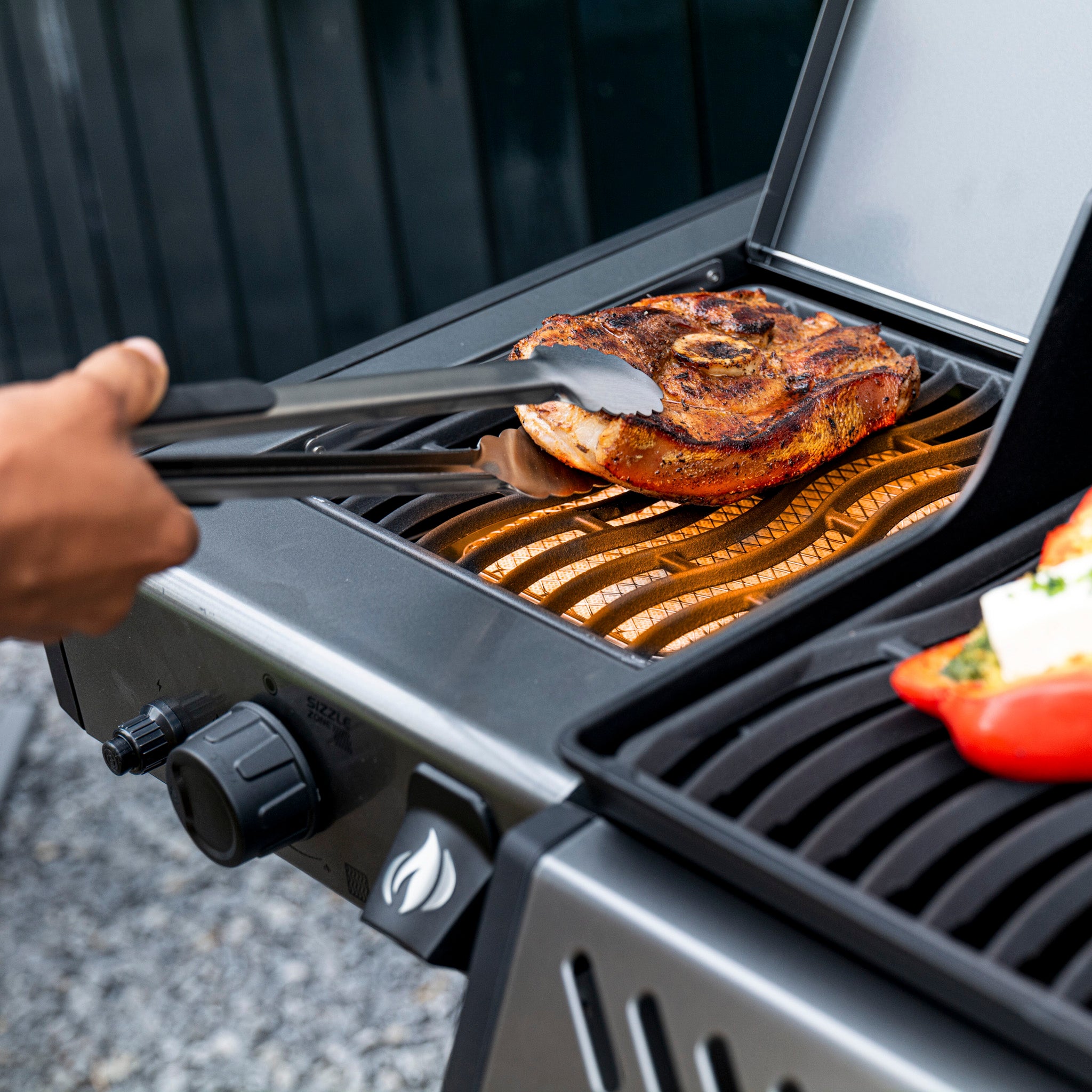 Person grilling a large piece of meat on a Napoleon Freestyle 365 SIB Gas Barbecue, using tongs, with a stuffed pepper on the side. The JETFIRE ignition system ensures everything starts smoothly.