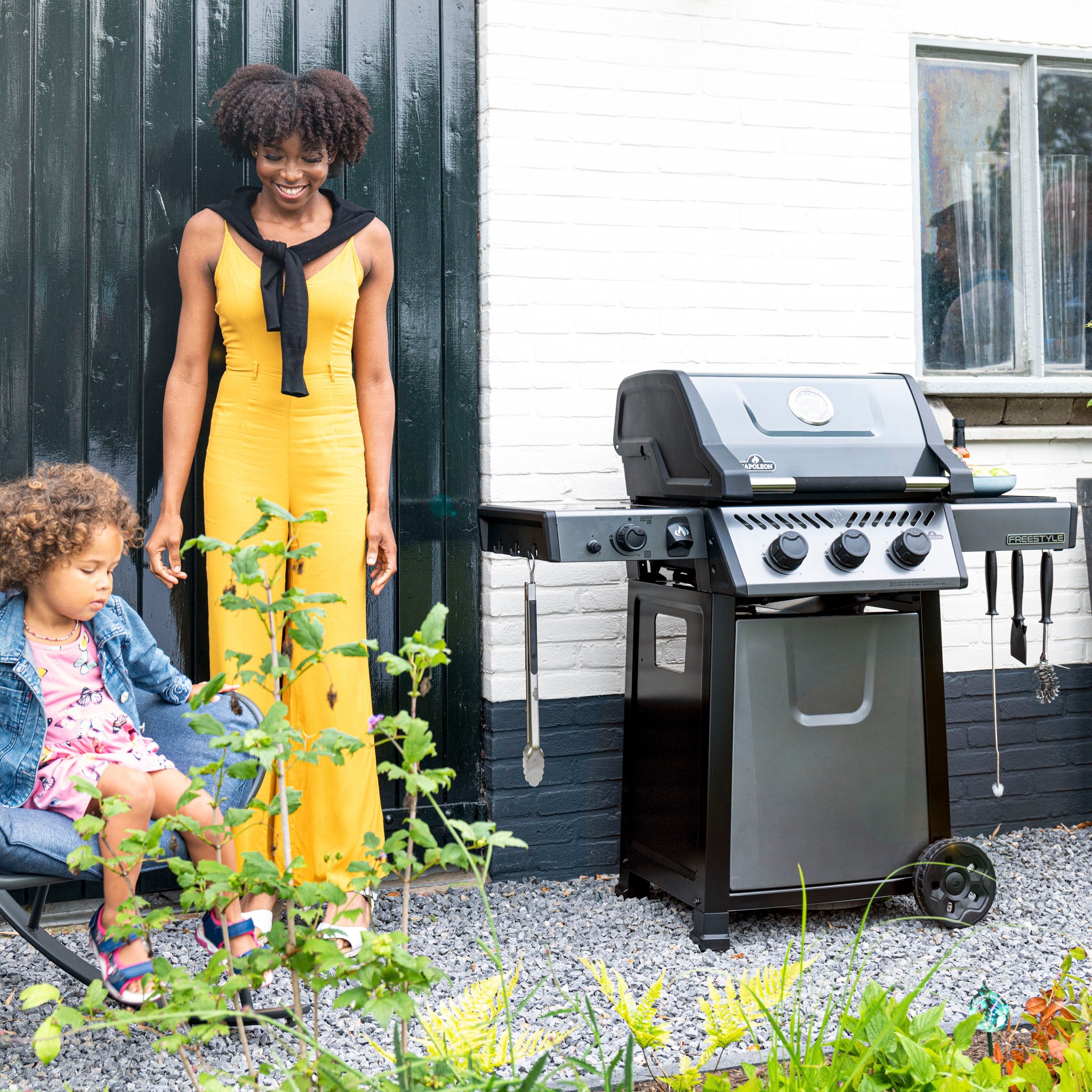 A woman in a yellow dress stands next to a Napoleon Freestyle 365 SIB Gas Barbecue, checking the JETFIRE ignition system, while a child sits nearby in the garden.