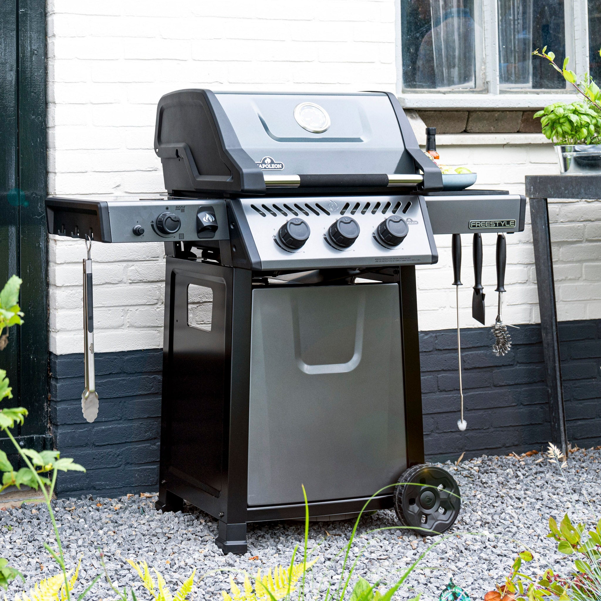 A Napoleon Freestyle 365 SIB Gas Barbecue, black in color, featuring four burners and an infrared side burner, complete with various utensils hanging on the sides, is placed outdoors on a gravel surface.