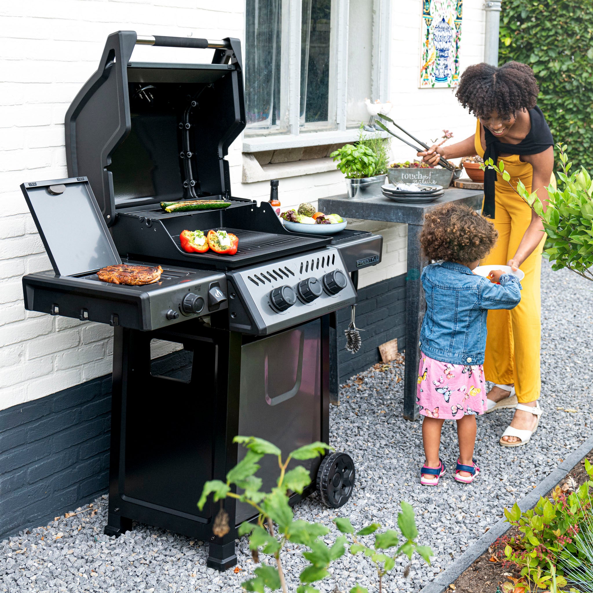 A woman assists a child near a Napoleon Freestyle 365 SIB Gas Barbecue, grilling vegetables and meat beside a house on a gravel path encircled by greenery.