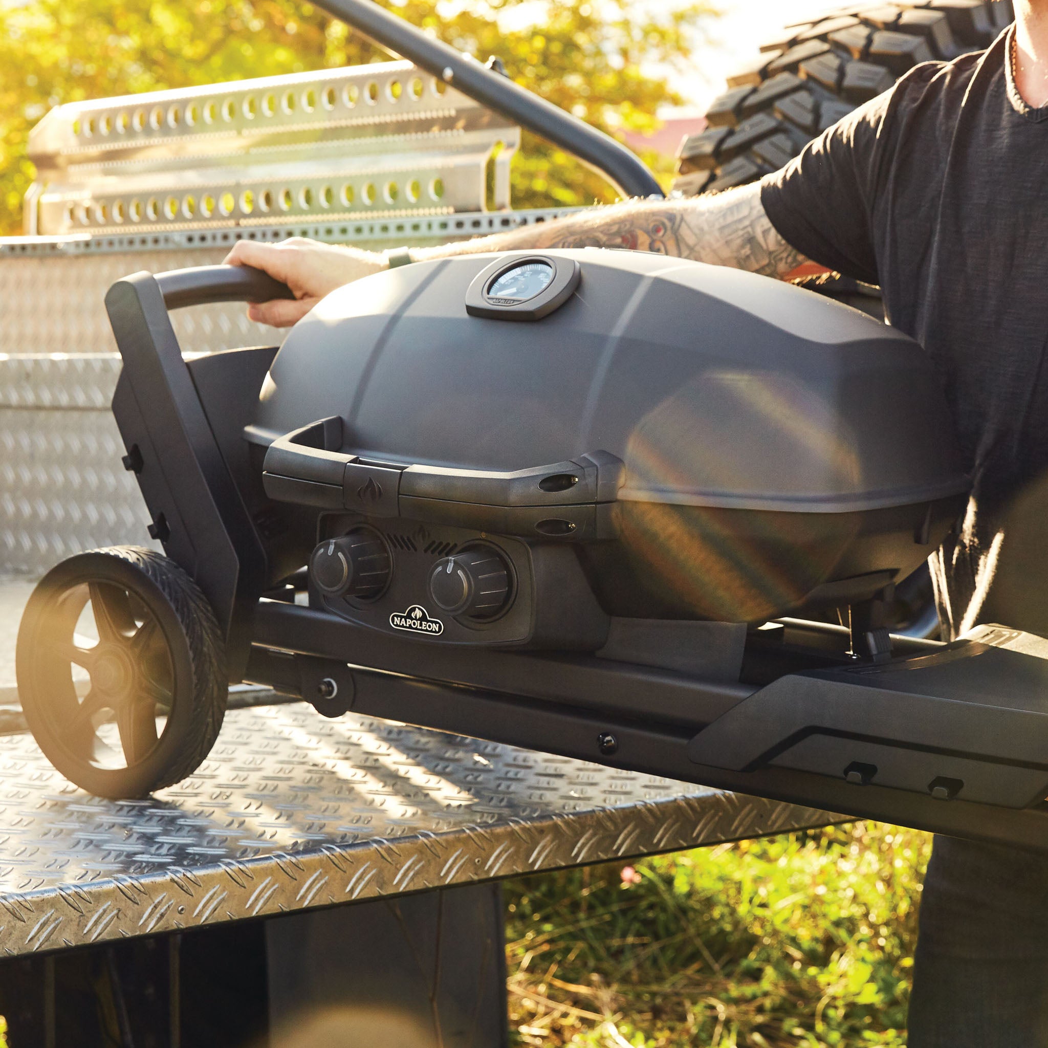 Person placing a Napoleon Phantom TravelQ Pro 285X Portable Gas Barbecue in black with wheels onto a metal step, with greenery and a tire in the background.