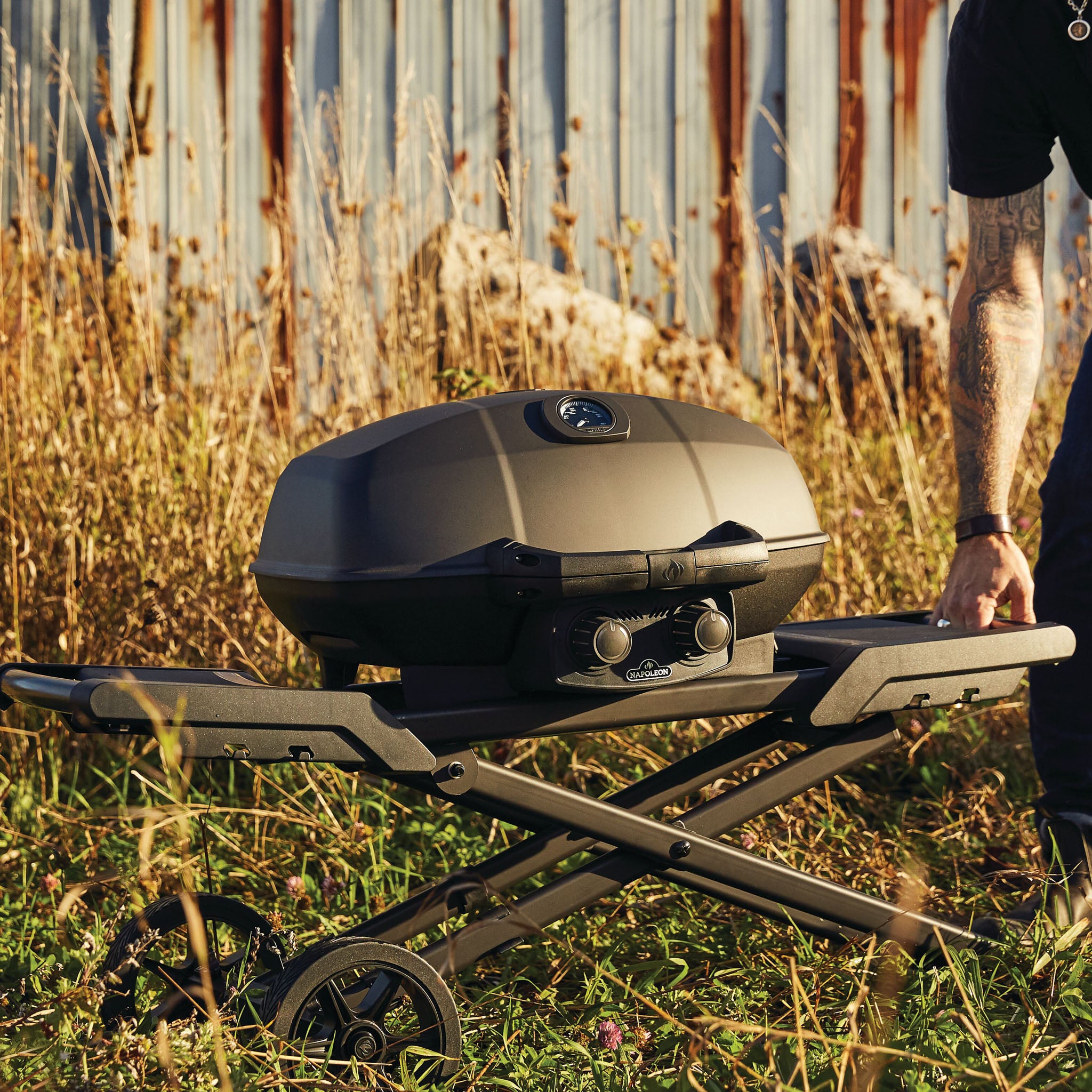 A person adjusts the Napoleon Phantom TravelQ Pro 285X Portable Gas Barbecue on a foldable stand in an overgrown grassy area.