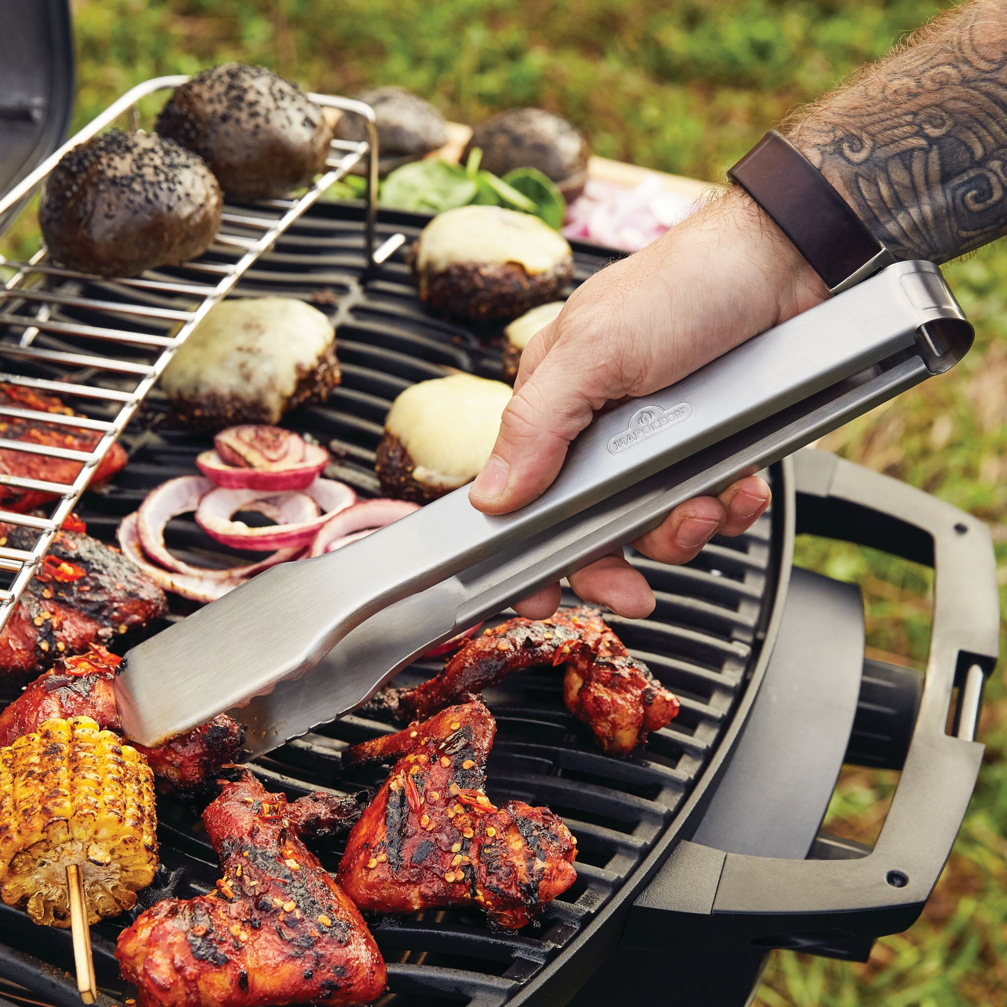 A person is using tongs to grill chicken, corn, and vegetables, including mushrooms and onions, on a Gourmet Portable Grilling setup featuring the Napoleon Phantom TravelQ Pro 285X Portable Gas Barbecue.