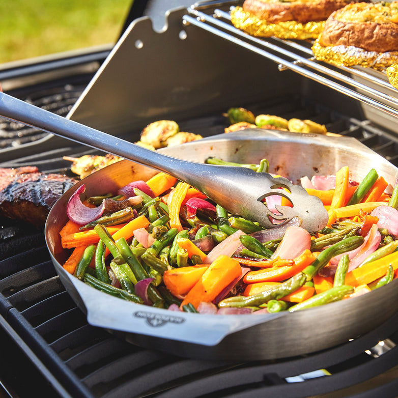 A skillet filled with colorful grilled vegetables being cooked on a Napoleon Rogue XT 365 SIB Gas Barbecue, with tongs reaching into the pan.