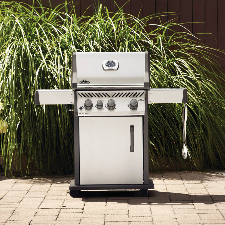 The Napoleon Rogue XT 425 SIB Gas Barbecue, a stainless steel propane gas grill with a closed lid, side shelves, and a hanging cloth, featuring an infrared side burner, is set on a patio with tall grass in the background.