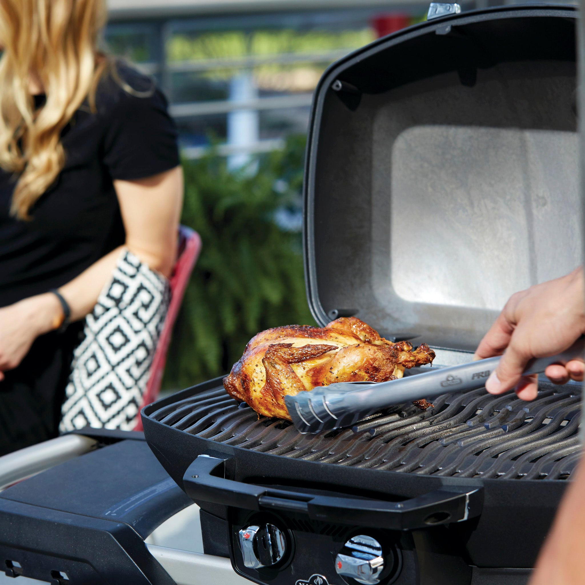 A person grilling a whole chicken on an outdoor BBQ grill, specifically the Napoleon Phantom TravelQ Pro 285X Portable Gas Barbecue, with another person sitting in the background.