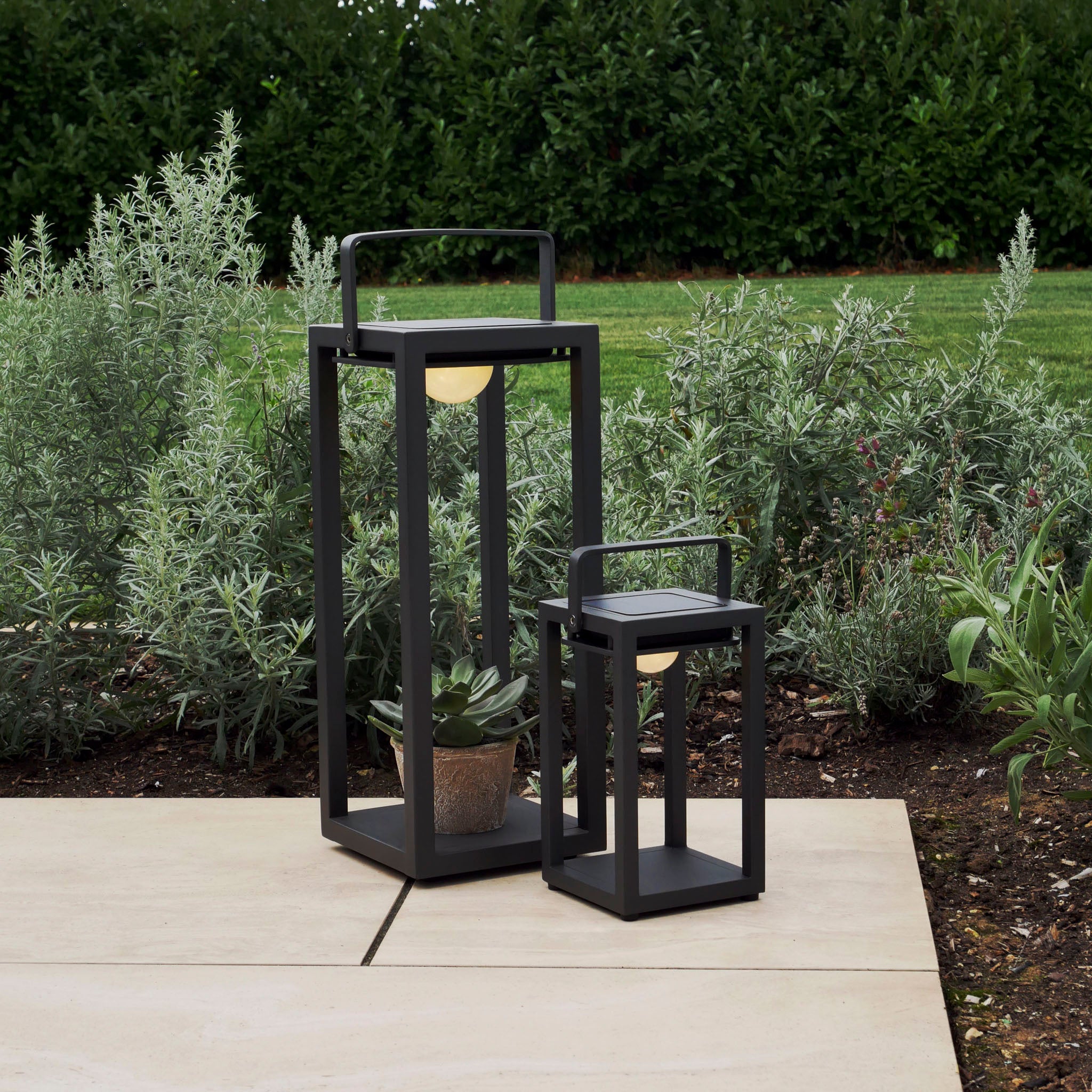 Two modern, weatherproof Luxor Small Table Outdoor and Indoor Solar Lanterns in Charcoal sit on a patio next to a garden with lush greenery.