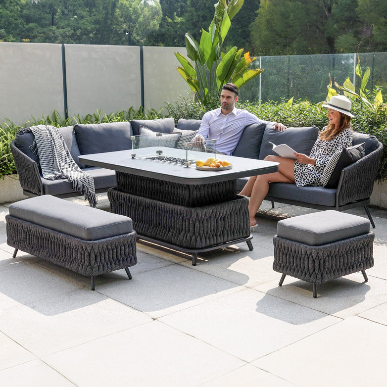 Two people relax on a Palma Rope Rectangular Corner Dining Set with Rising Firepit Table in Grey (Left Hand), featuring sturdy aluminum frames and comfortable gray cushions; the table displays an assortment of fruit, surrounded by lush greenery and modern decor.