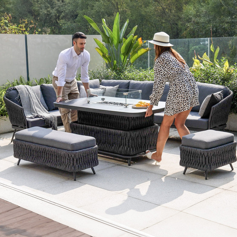 Two people set a table in a modern outdoor patio adorned with the Palma Rope Rectangular Corner Dining Set with Rising Firepit Table in Grey (Left Hand). The comfortable seating, featuring aluminium frames and a hydrophobic thick rope weave, is complemented by lush greenery in the background.