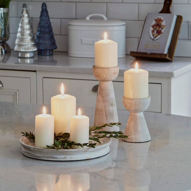 Lit white candles elegantly rest on the kitchen counter in Ivyline's Padstow Whitewash Wooden Candle Holders, surrounded by lush greenery. Decorative trees and a cookbook in the background add charm, enhancing the cozy ambiance.