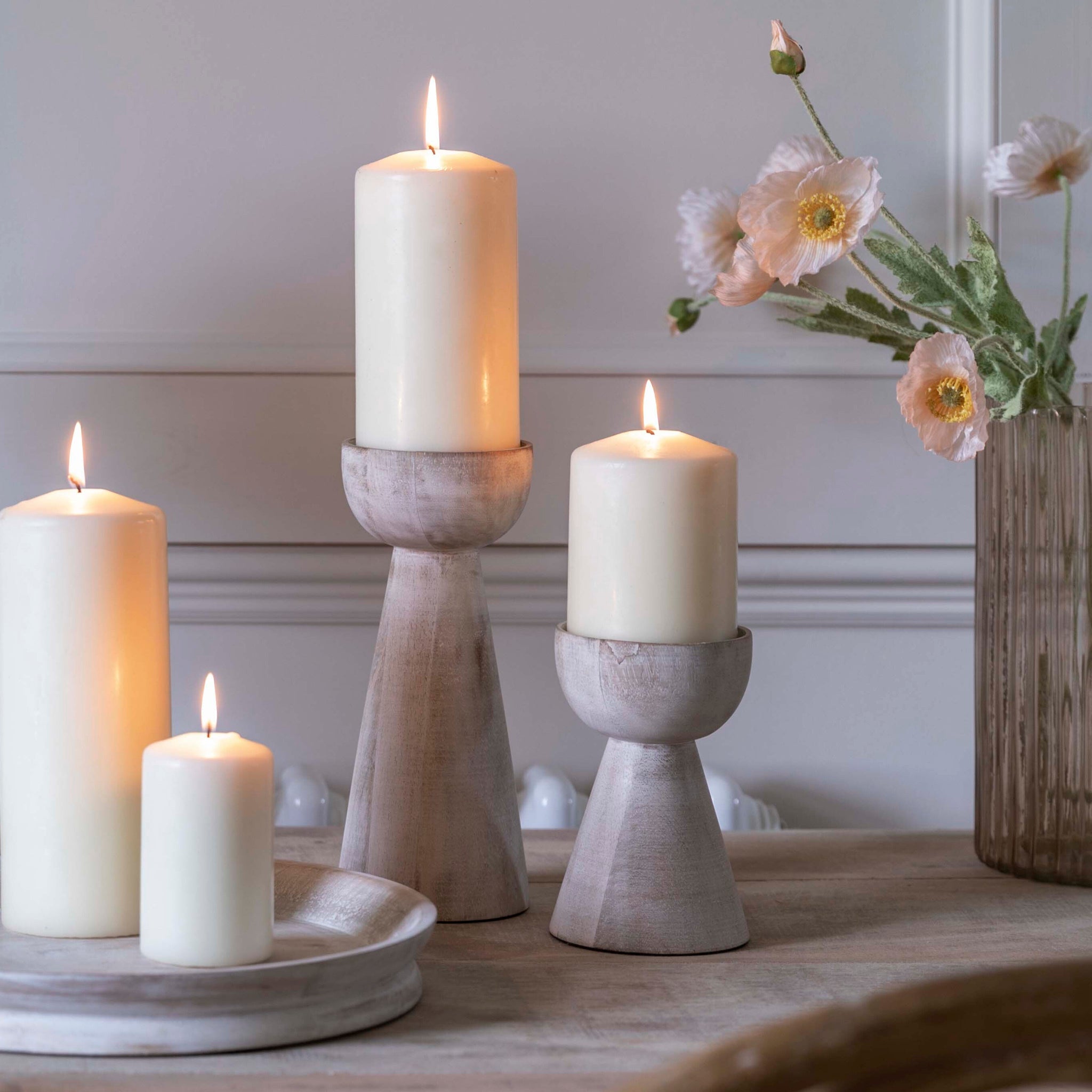 White candles flicker warmly in Ivyline's Padstow Whitewash Wooden Candle Holders on the table. Nearby, white flowers rest elegantly in a ribbed glass vase, creating a serene ambiance.